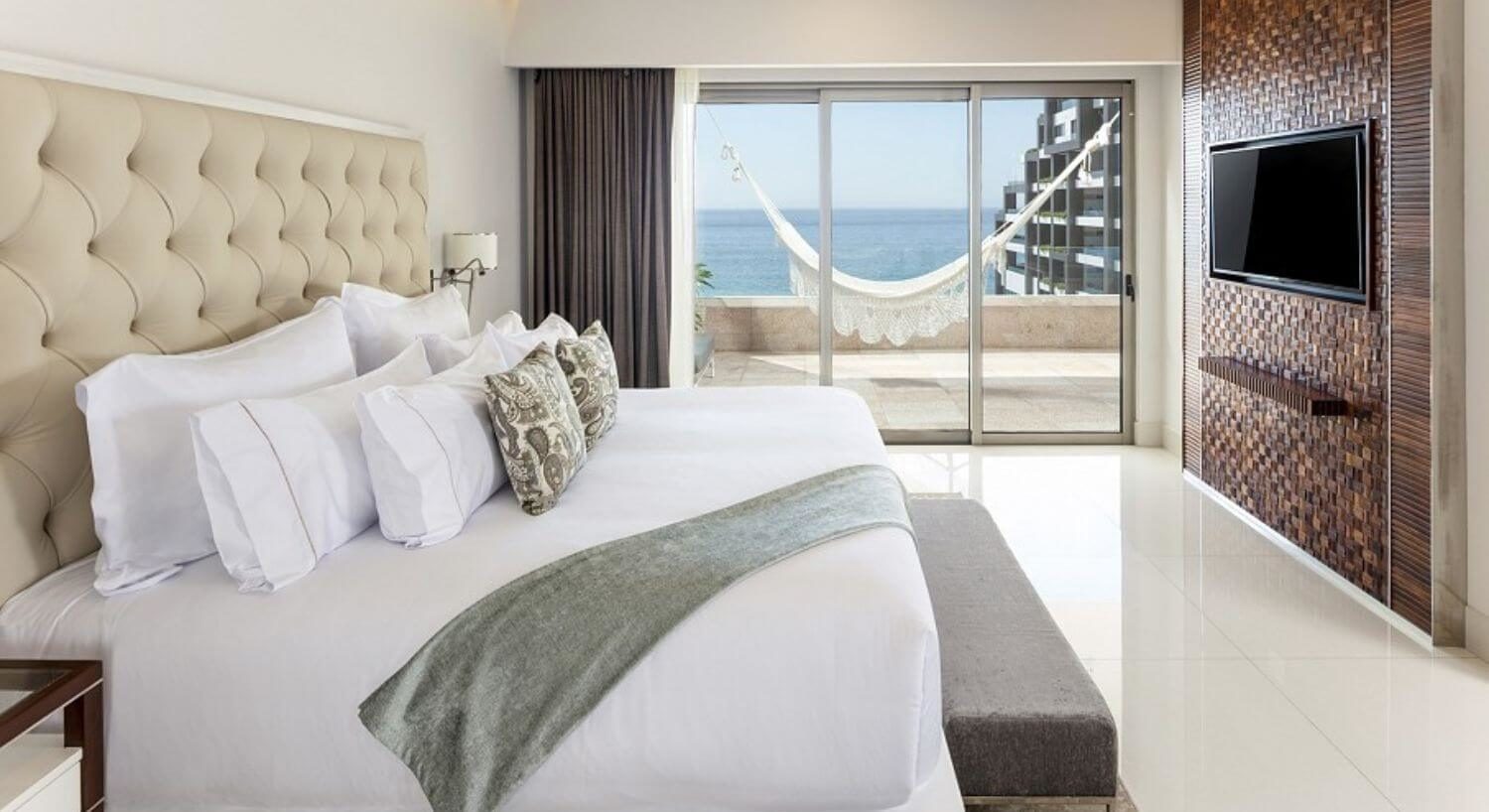 A bedroom with a king bed with plush headboard, a sitting bench at the foot of the bed, a flat screen TV on the opposite wall, and sliding doors leading out to a private balcony with patio furniture, hammock, and ocean views.