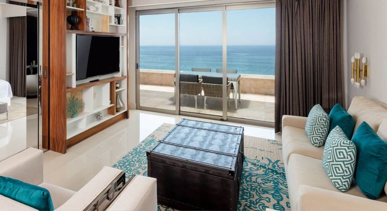 Chic upscale living room with white furniture, a wood chest, an entertainment center with flat screen TV, a bed in the bedroom, and sliding doors in the living room opening to a patio with furniture and ocean views.
