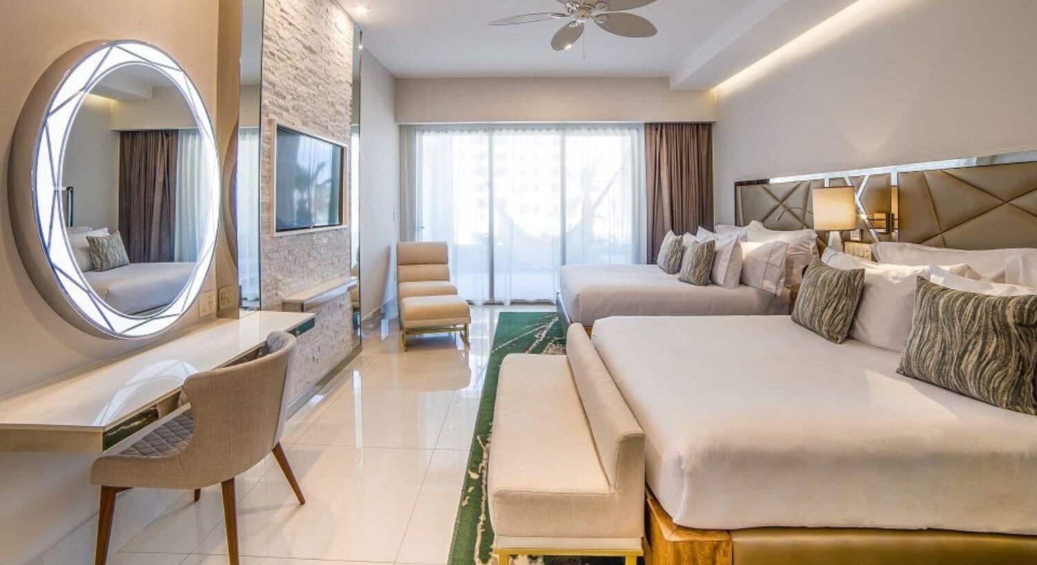 A bedroom with 2 queen beds, a sitting bench at the foot of one bed, a writing desk and chair with mirror on the opposite wall, along with a flat screen TV and chaise lounge, and sliding doors that lead out to a private balcony with patio furniture and hammock.