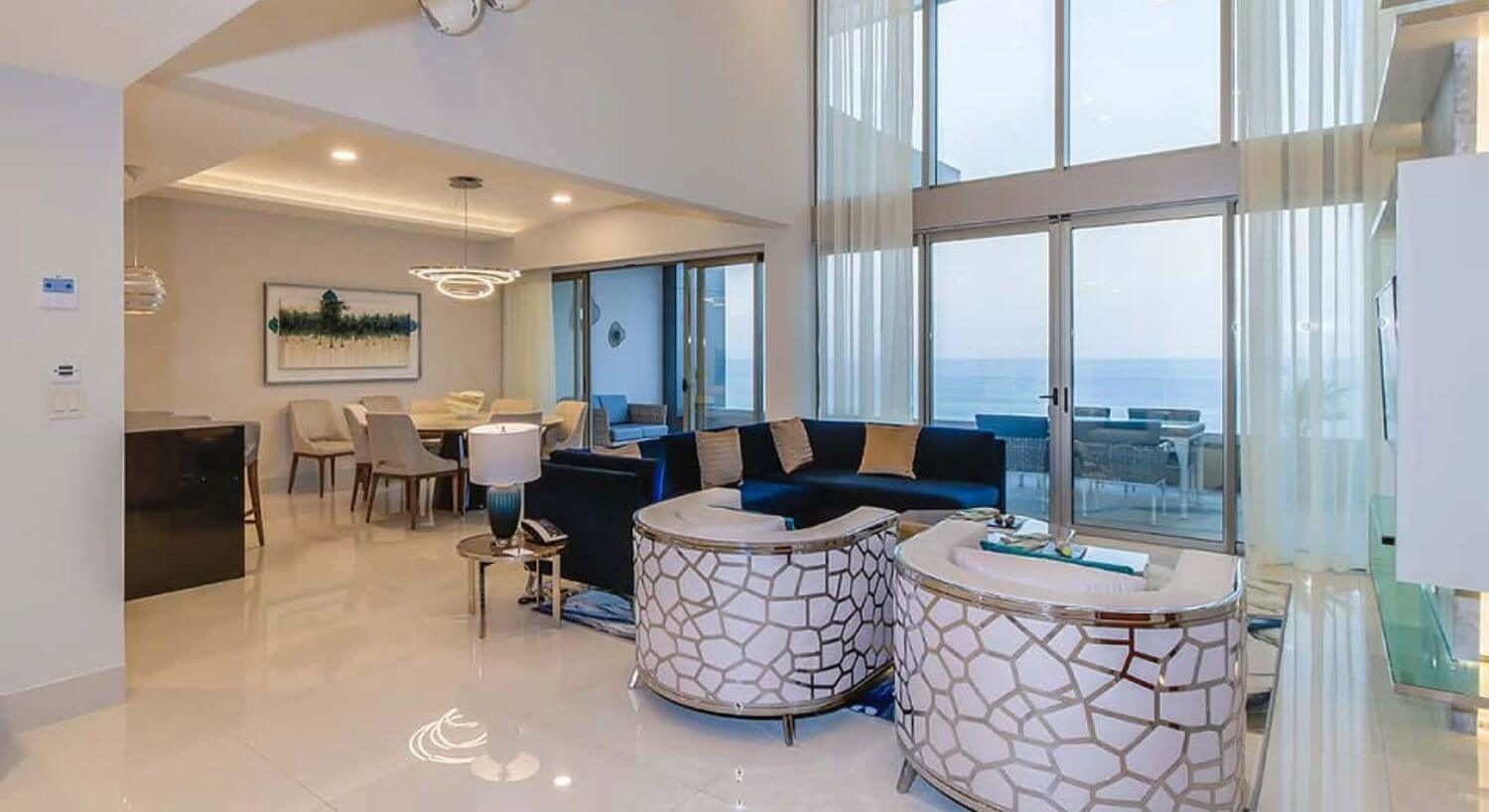 A large open floor plan living room with multi story ceilings, adining area with blue sofas, plush white armchairs, nightstands with lamps, a flat screen TV, a round dining table and 8 chairs, and sliding doors leading out to a private balcony with patio furniture and ocean views.