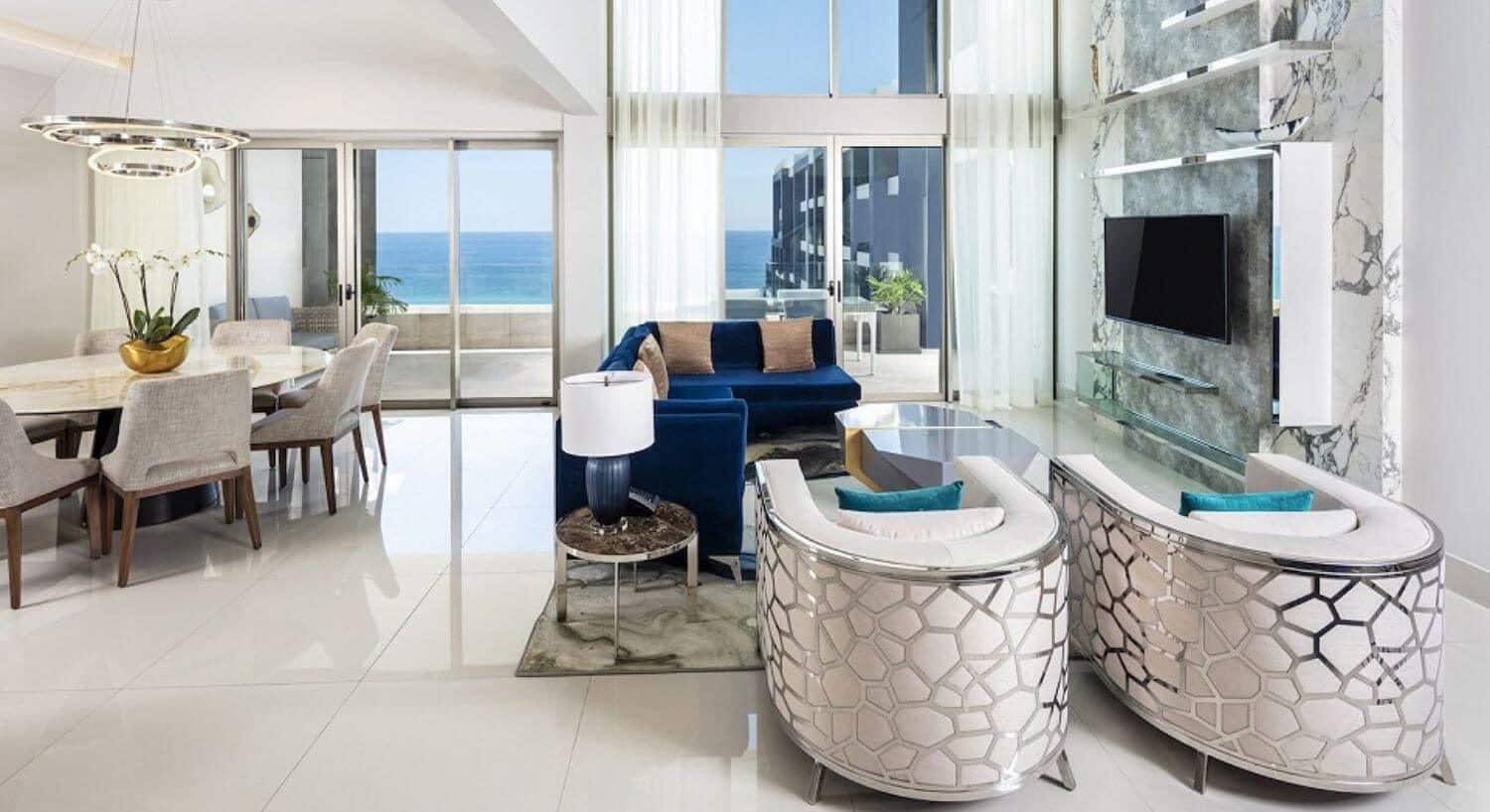 A large open floor plan living room and dining area with blue sofas, plush white armchairs, nightstands with lamps, a flat screen TV, a round dining table and 8 chairs, and sliding doors leading out to a private balcony with patio furniture and ocean views.