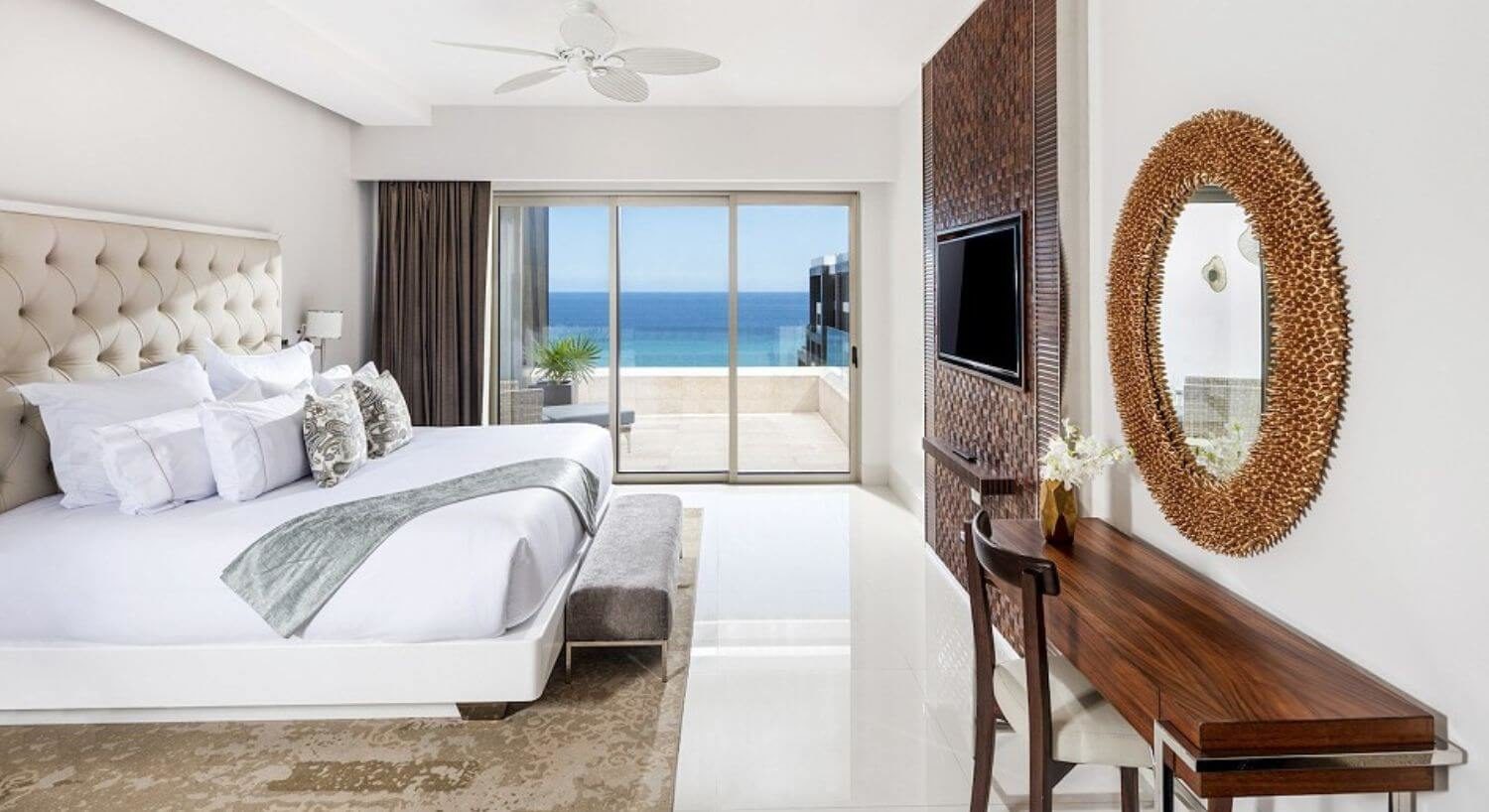 A bedroom with a King bed with plush headboard and a sitting bench at the foot of the bed, a flat screen TV and writing desk and chair on the opposite wall, and sliding glass doors that lead out to a private balcony with patio furniture and ocean views.