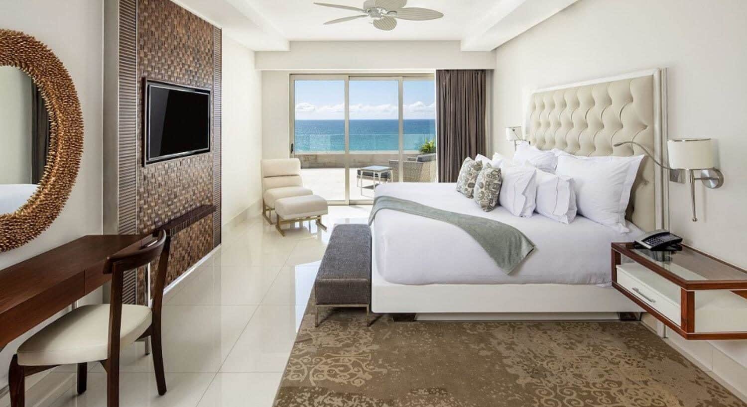 A bedroom with a King bed, a plush headboard, a sitting bench at the foot of the bed, a flat screen TV and writing desk and chair on the opposite wall, a chaise lounge chair in the corner, and sliding doors that lead out to a private balcony with patio furniture and ocean views.
