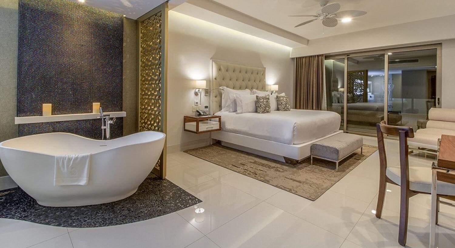 A bedroom with a king bed, nightstands with lamps on either side, a writing desk and lounge chair on the opposite wall, a deep soaking tub, and sliding glass doors that lead out to a private balcony.
