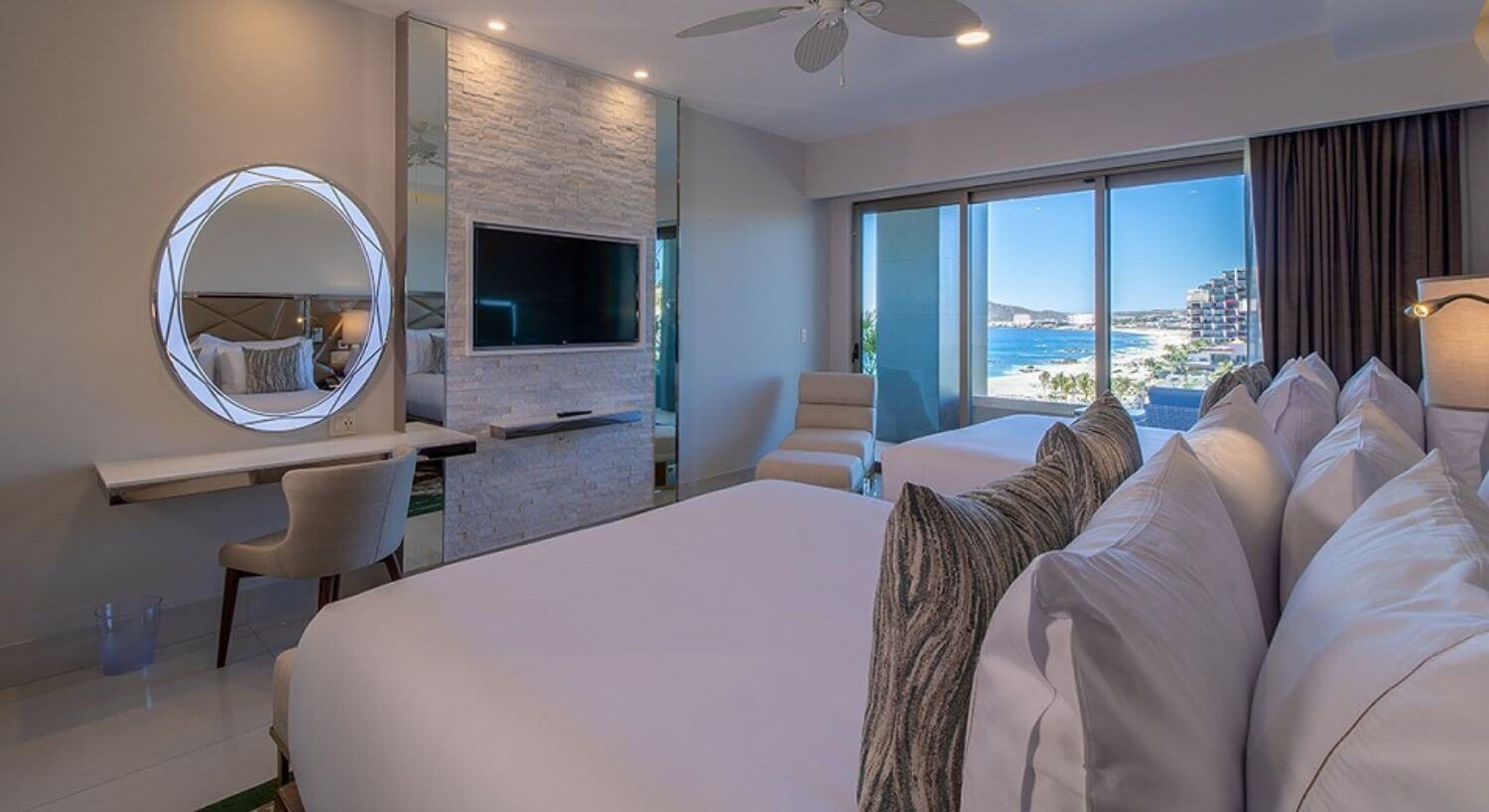A bedroom with 2 queen beds, a flat screen TV and writing desk and chair on the opposite wall, a chaise lounge chair in the corner, and sliding doors out to a private balcony with patio furniture, and the beach and ocean views.