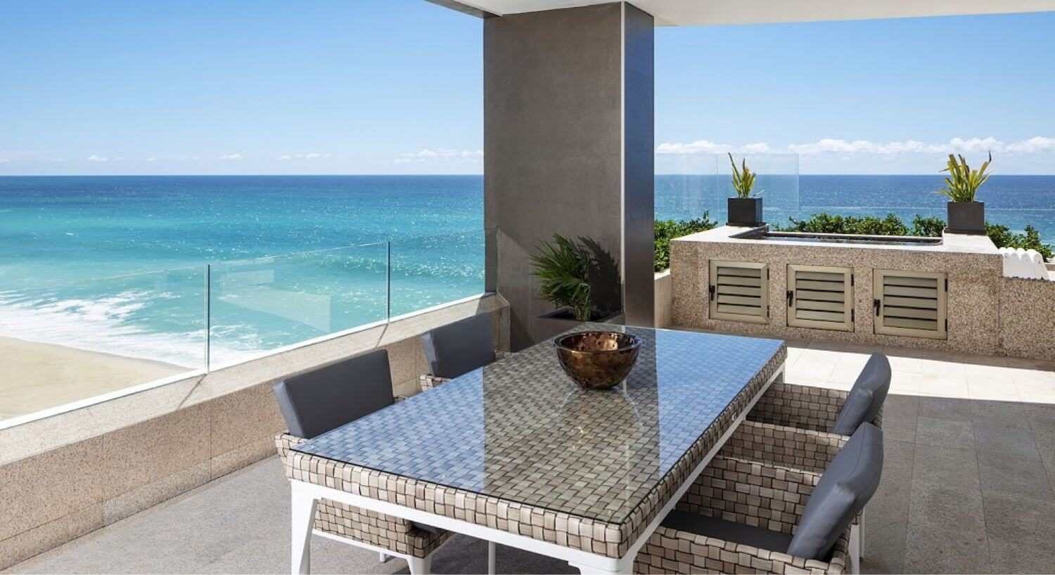 A balcony with a dining table and chairs, a jacuzzi, and views of the sandy beach and turquoise and blue ocean.