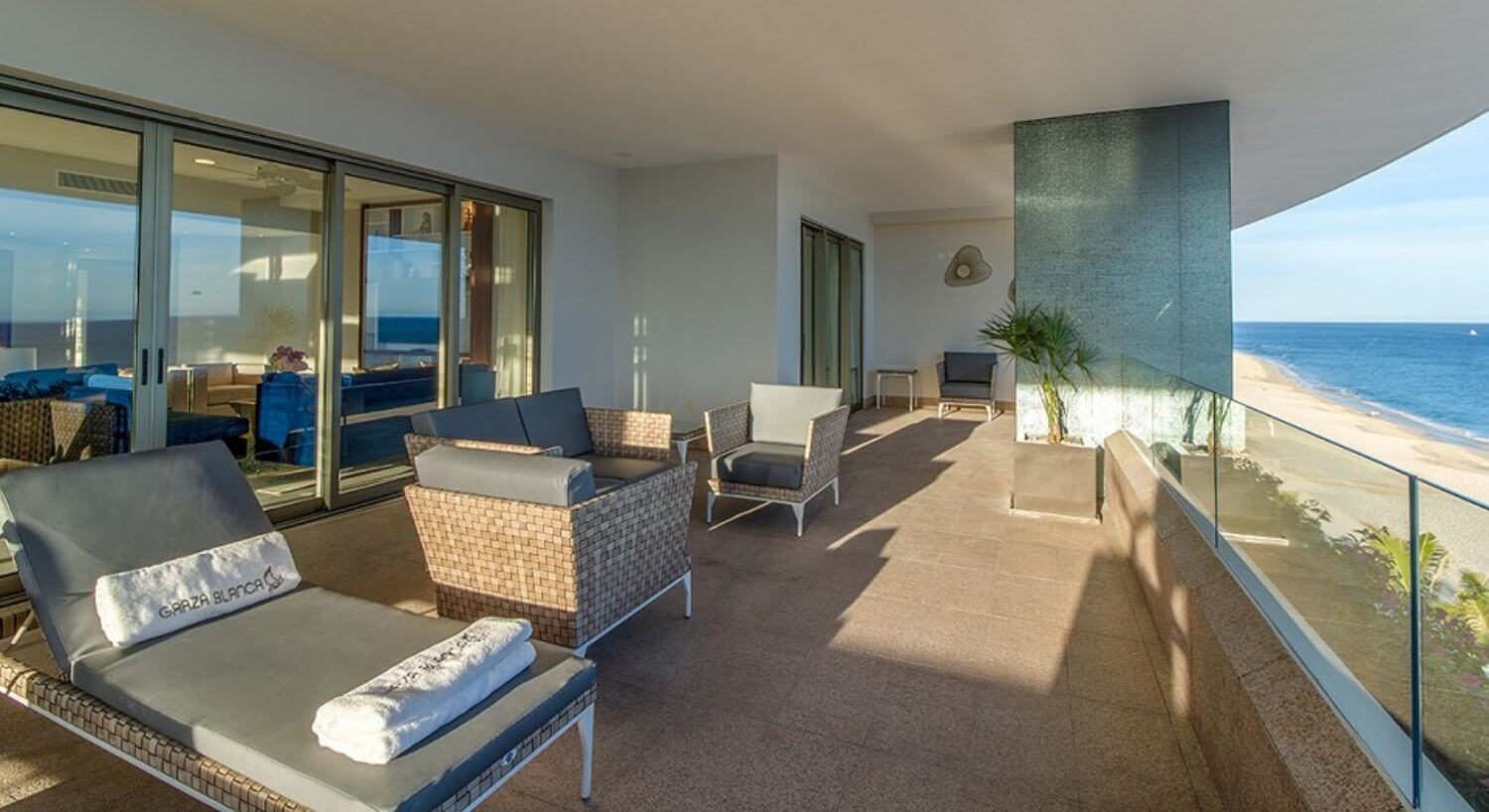 A spacious balcony with patio furniture, lounge chairs, sliding doors open doors into a living room, and views of the sandy beach and ocean.