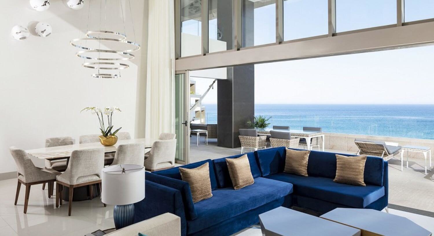A large living room and dining area with 2 story ceilings and open sliding doors that span the width of the room and open out to a large balcony with outdoor patio furniture and sweeping ocean views.