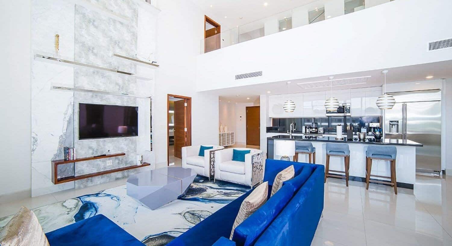 An open floor plan living room and kitchen with blue sofas and white armchairs, a flat screen TV, a gourmet kitchen with stainless steel appliances, and a door open to a bathroom.