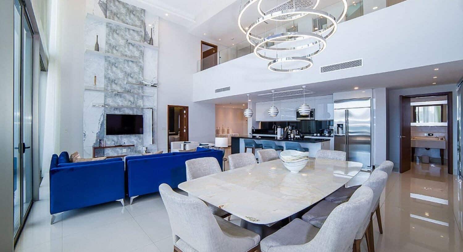 A large multi story open floor plan living room with blue sofas and white armchairs, a dining area with table and 8 chairs, a gourmet kitchen, a door open to a bathroom, and a second story with open doors to various rooms.
