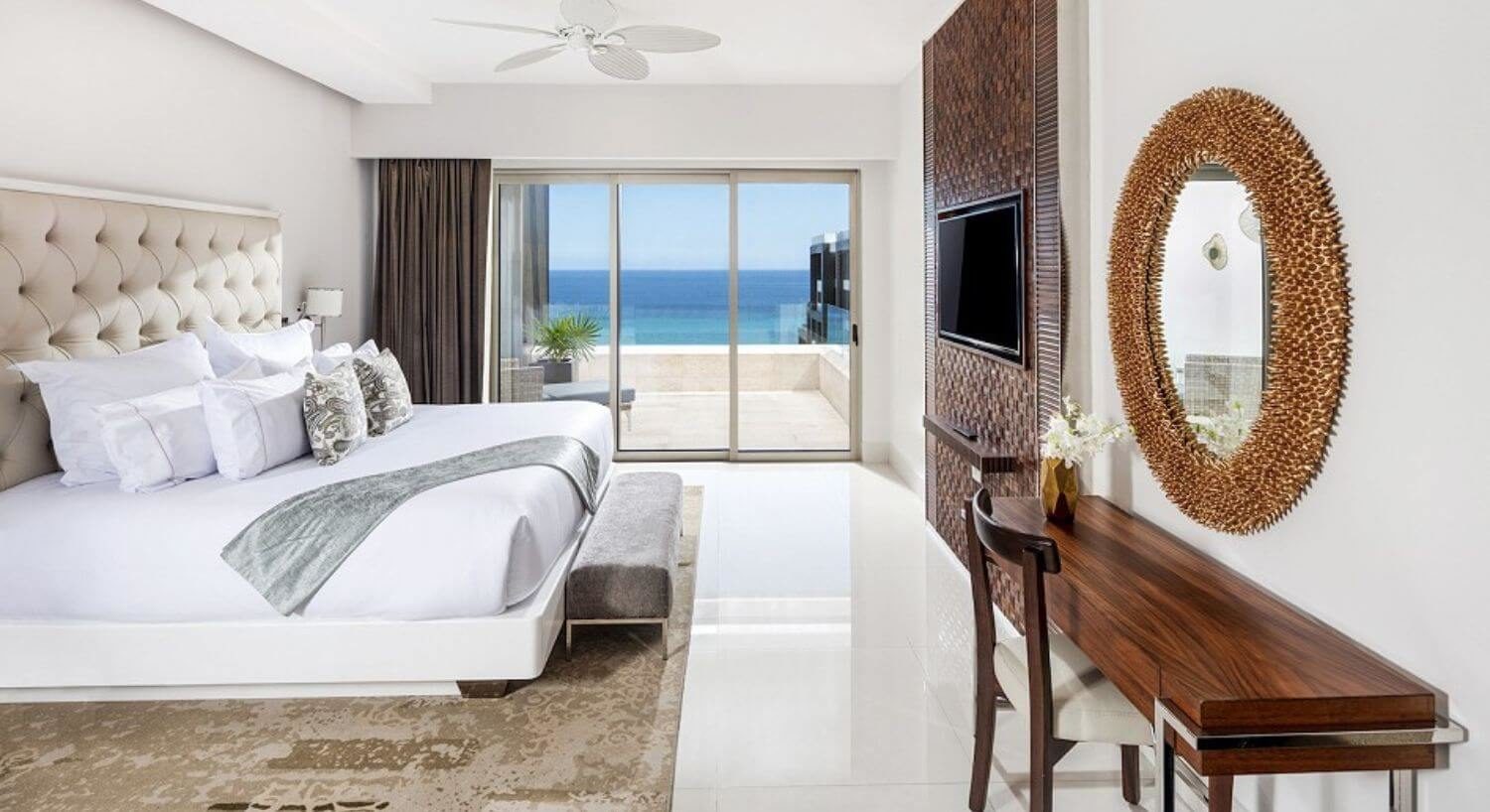 A bedroom with a King bed, plush headboard, sitting bench at the foot of the bed, a writing desk and chair with mirror above it, a flat screen TV on the wall opposite the bed, and sliding doors out to a private balcony with patio furniture and ocean views.