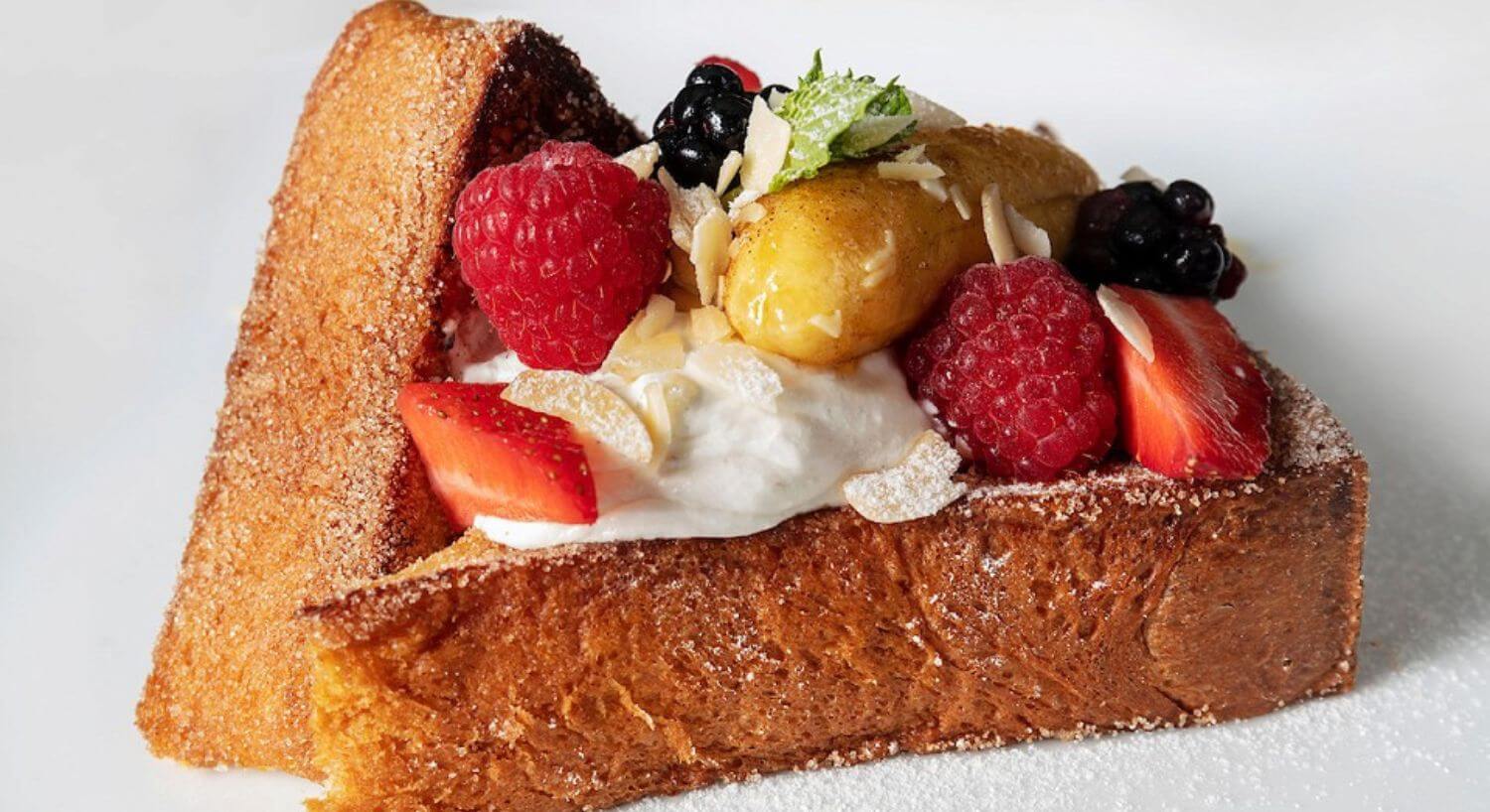 Two triangular pieces of french toast with a dollop of cream, topped with fresh berries, mint sprigs, and powdered sugar.
