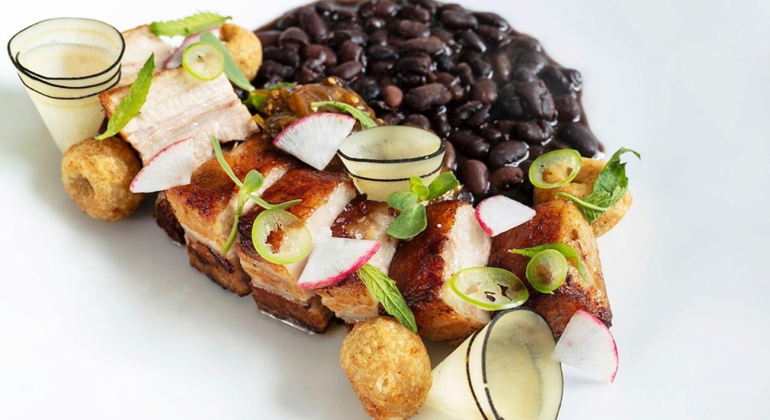 A white plate with black beans, fried pork belly pieces, and garnished with pieces of fresh sliced vegetables.