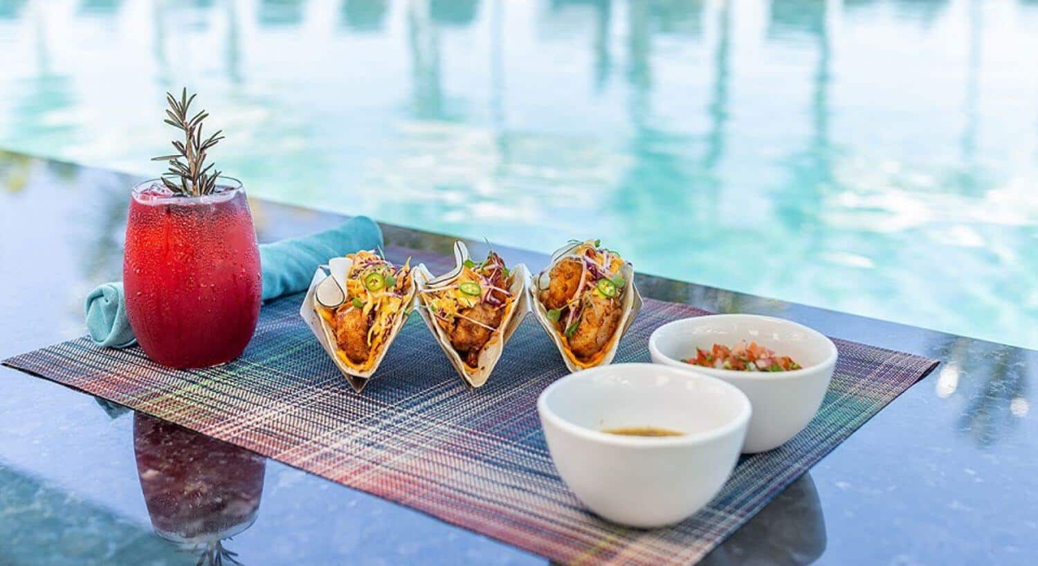 A trio of fish tacos on a colorful placemat with bowls of salsa on the side, a turquoise napkin, and a red cocktail with a sprig of fresh rosemary, in front of a glimmering pool.