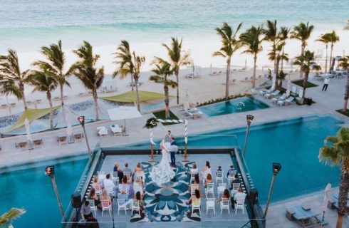 A wedding with a bride and groom saying their vows in front of a number of people on white chairs on a platform over a pool with the beach, palm trees, and the ocean in the background.