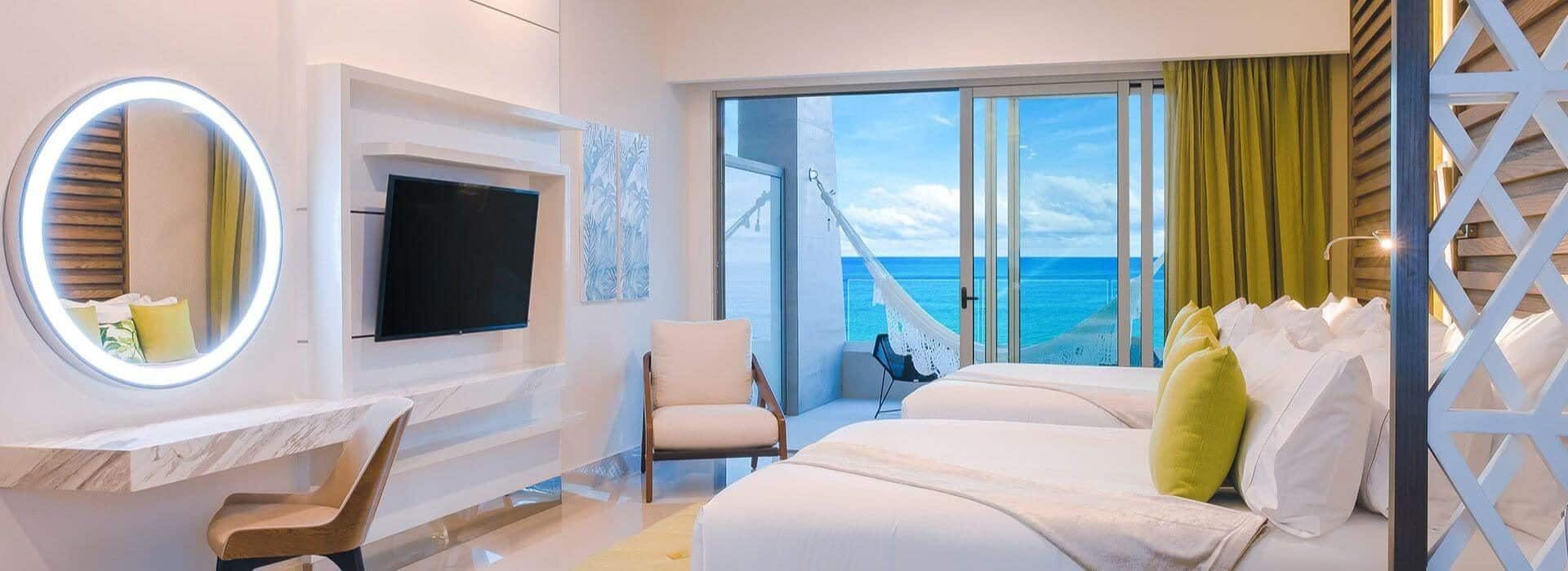 Bedroom with 2 beds with white linens, green accent pillows and throws, a writing desk with lighted circular mirror above it, a flat screen TV, and sliding glass doors leading out to a patio with ocean views