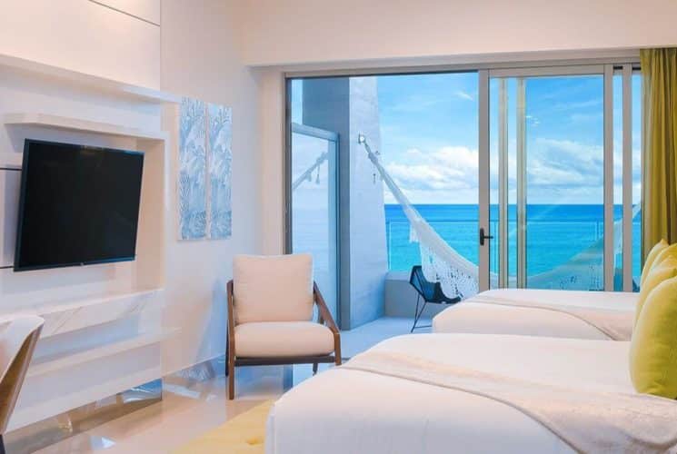 Bedroom with 2 beds with white linens, green accent pillows and throws, a writing desk with lighted circular mirror above it, a flat screen TV, and sliding glass doors leading out to a patio with ocean views
