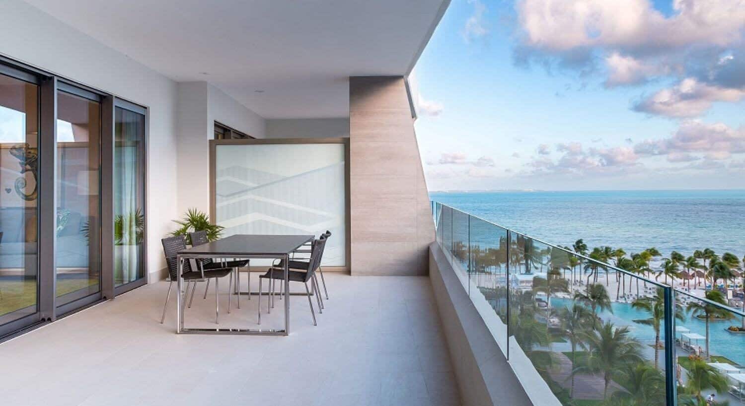A private balcony with a dining table and chairs, sliding doors into a living room, and views of the resort pools, palm trees, sandy beach and ocean.