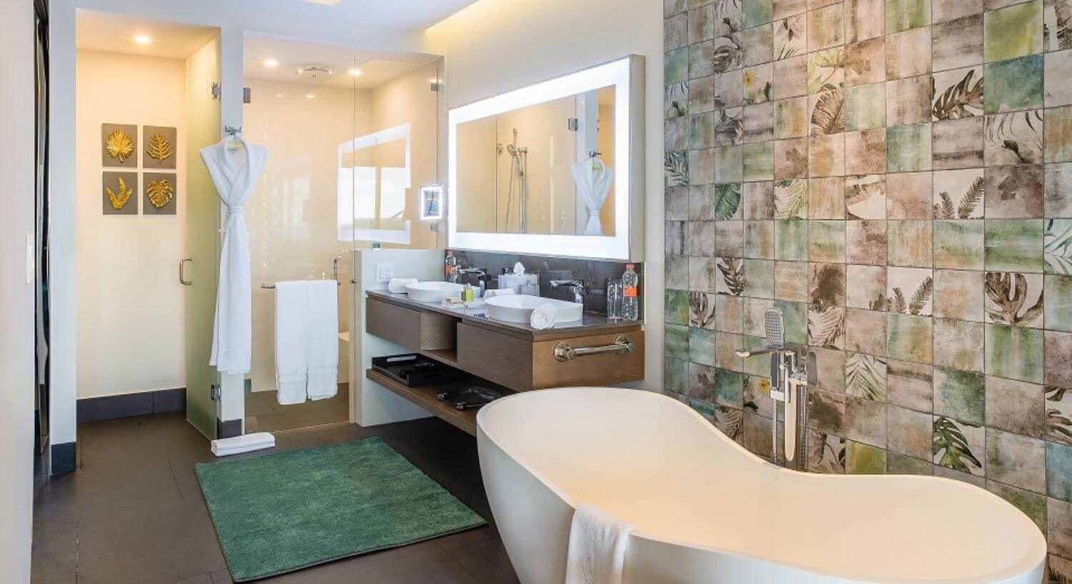 A bathroom with a deep soaking tub, a large vanity with 2 sinks and a lit mirror the width of the vanity above the sinks, along with a walk in shower.