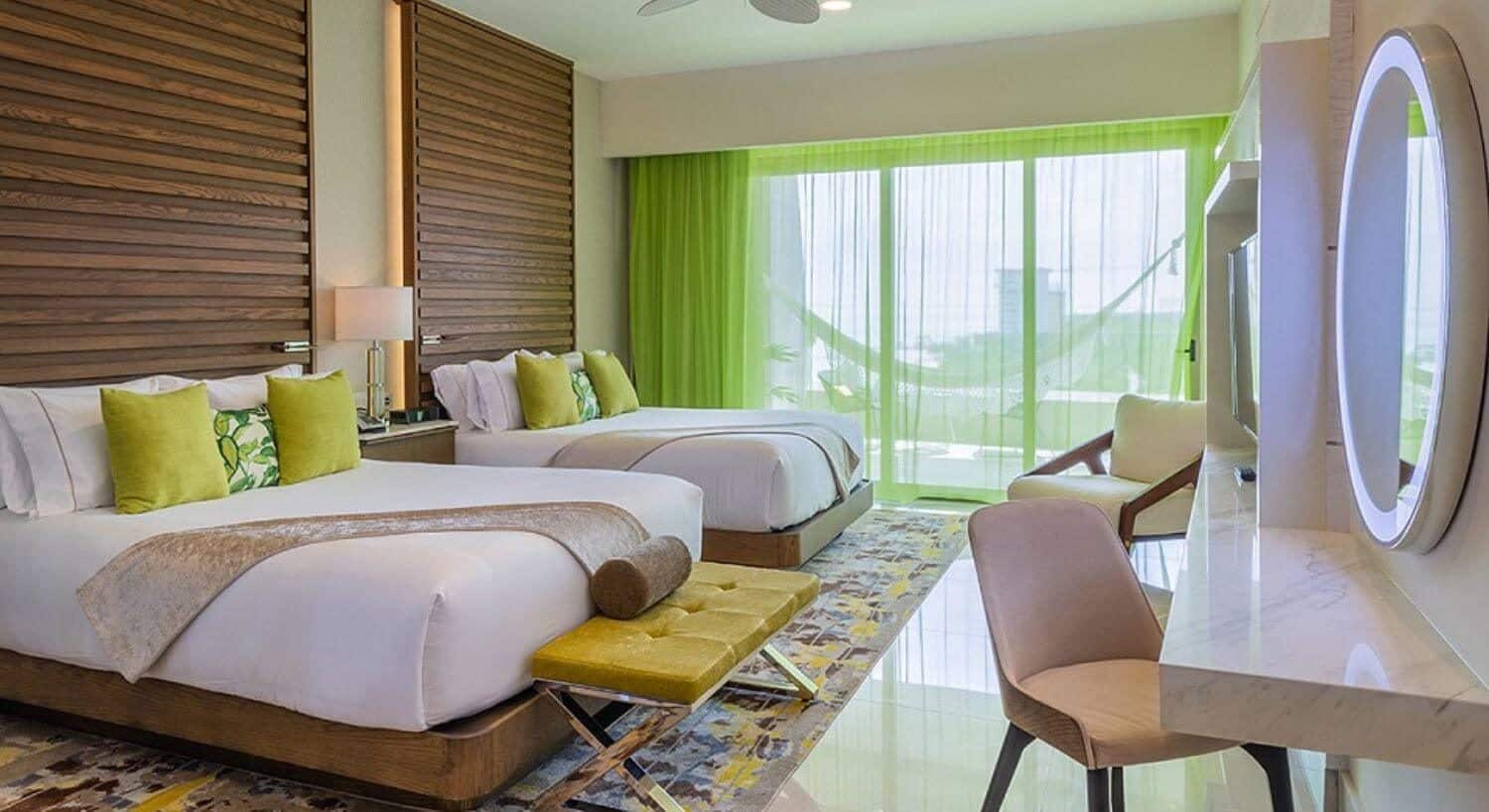 A bedroom with 2 queen beds with floor to ceiling wood headboards, a nightstand and lamp in between them, a bench at the foot of one bed, a writing desk and chair, and sliding doors with sheer green curtains that lead out to a private balcony with hammock.