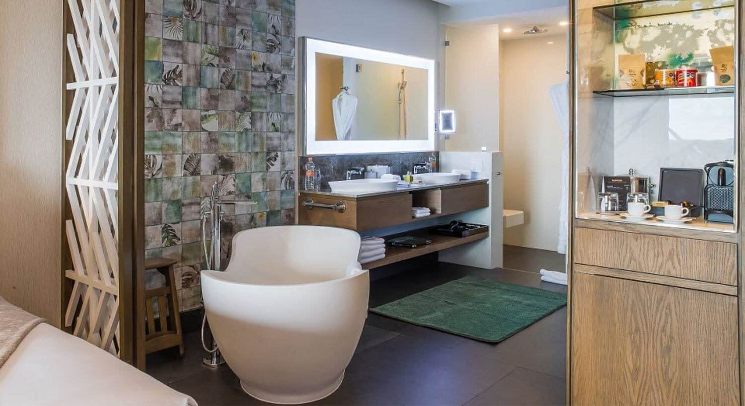 A bathroom with 2 sinks, a deep soaking tub, a separate shower, and a coffee and snack center near the bathroom, and a corner of a bed next to the bathroom.