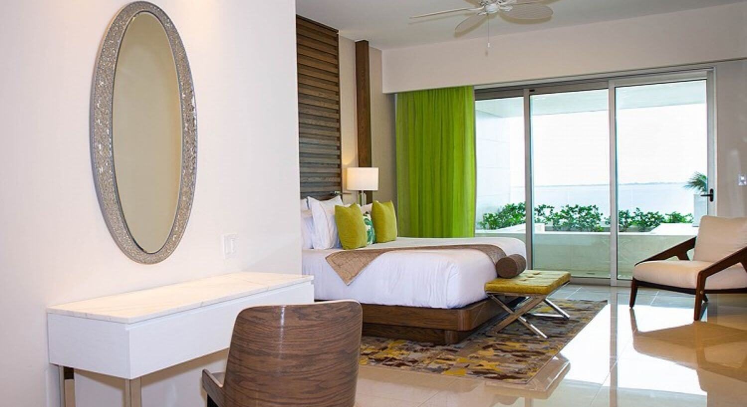 A bedroom with a King bed with white and green bedding, a bench at the foot of the bed, a writing desk and chair with a mirror above it, an armchair in the corner, and sliding glass doors leading out to a balcony with a hot tub and ocean views.