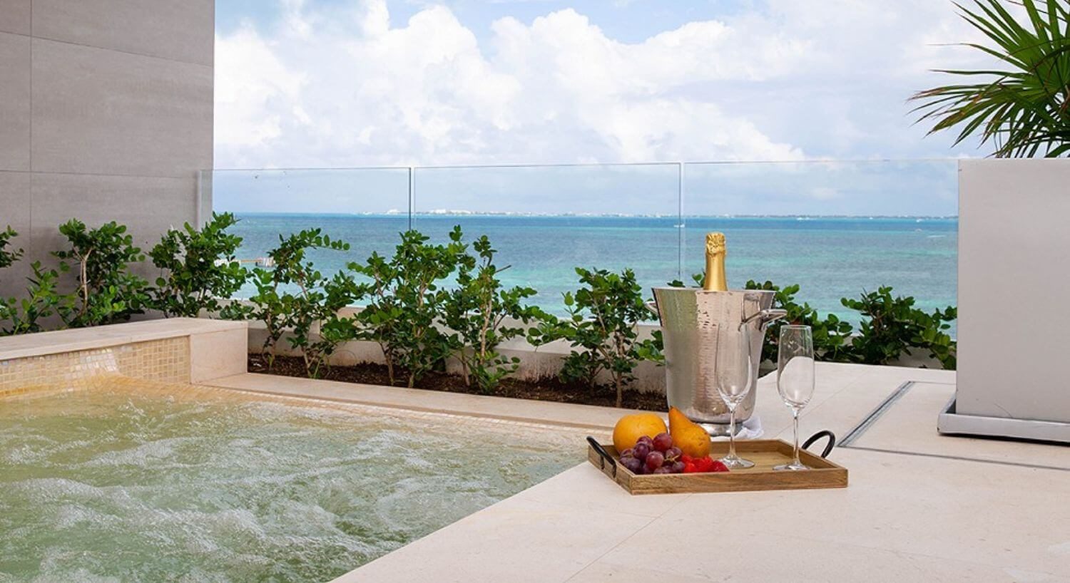 An outdoor hot tub bubbling away, with a wood platter of fruit and champagne glasses, along with a silver bucket of champagne, green bushes behind the hot tub, and turquoise ocean views.