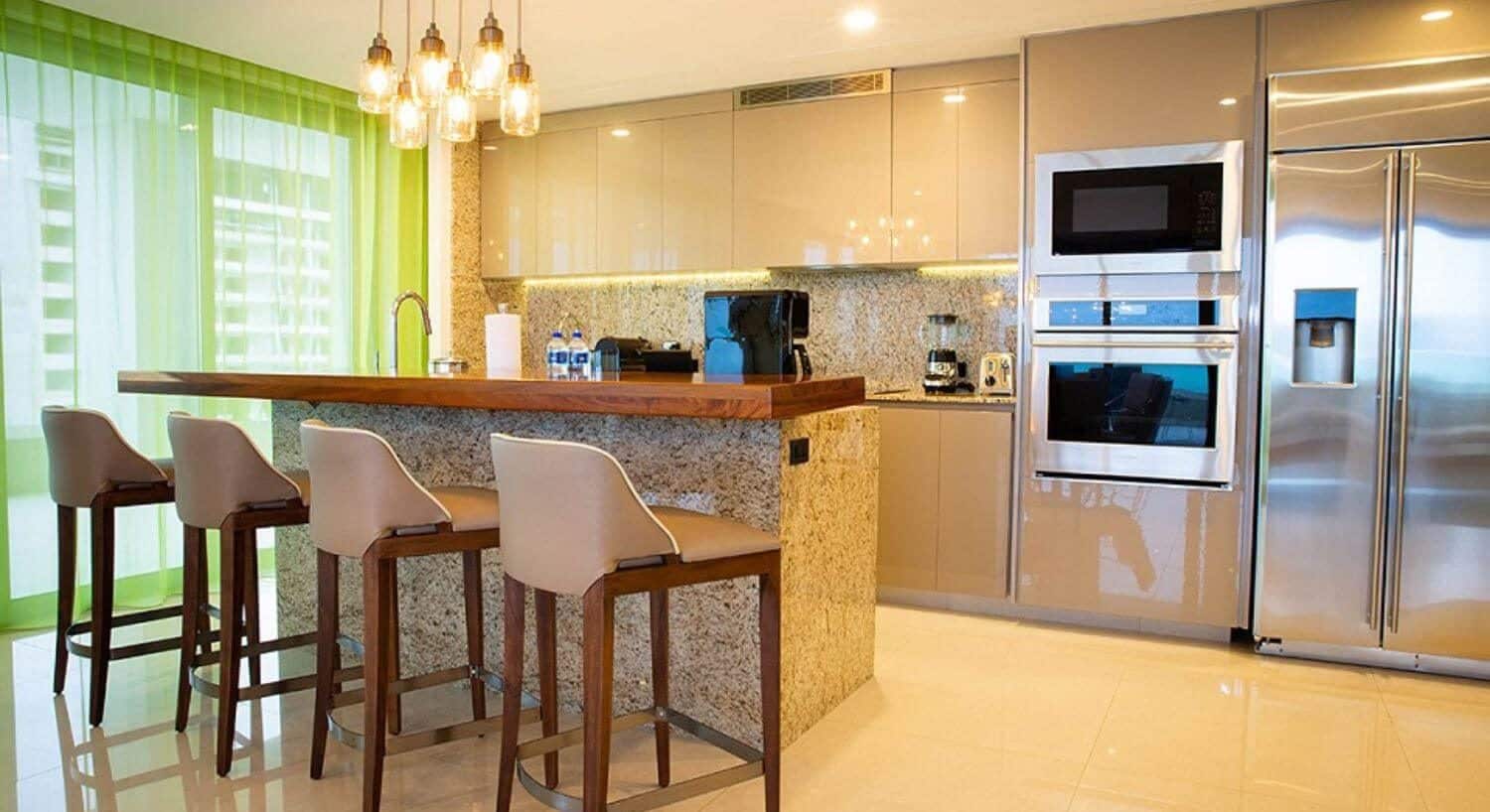 A gourmet kitchen with stainless steel appliances, a breakfast bar with 4 bar stools, and floor to ceiling windows with sheer green curtains that open out to a private balcony.