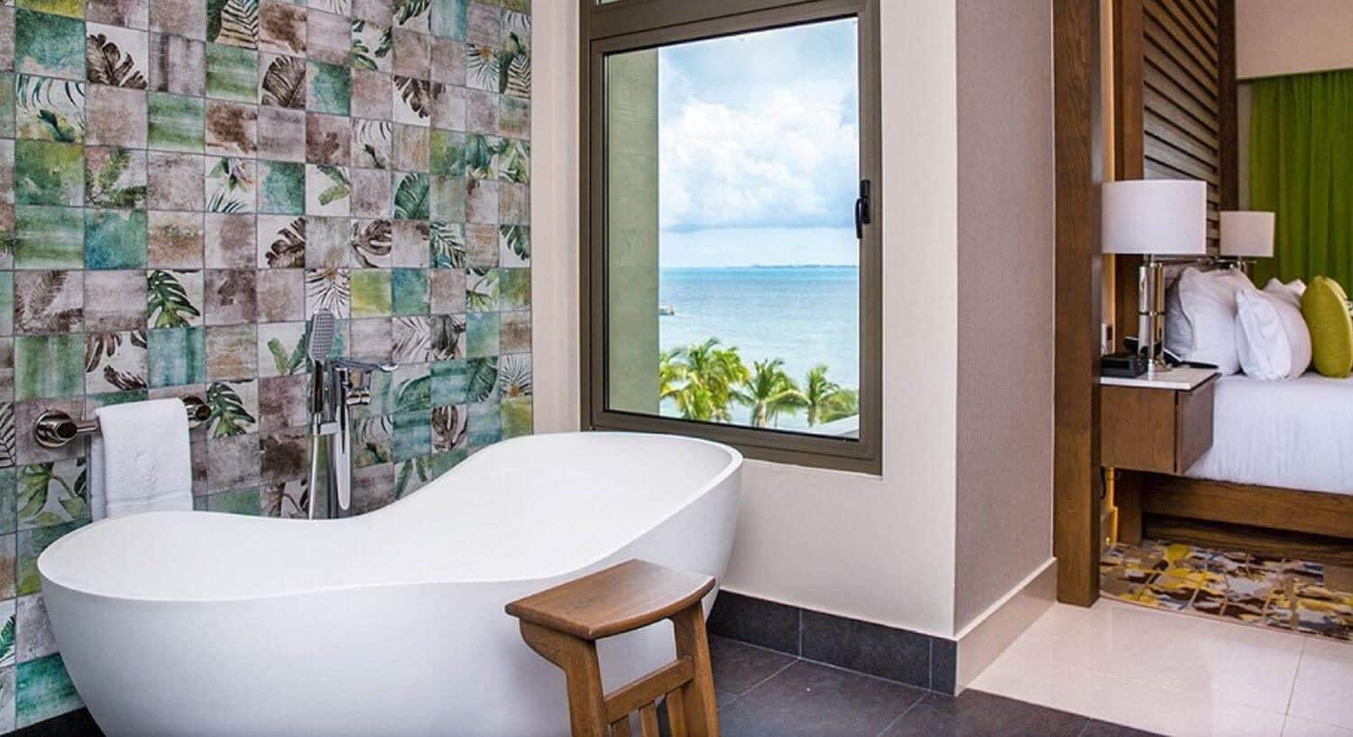 A deep white soaking tub with a window with ocean views and the tops of palm trees, and a bedroom with the head of the bed and nightstands.