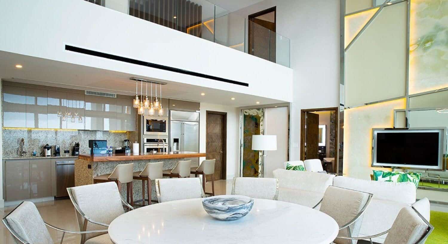 A multi story room with a dining area with white table and chairs, a living area with white sofa and chairs, a large flat screen TV, a gourmet kitchen with stainless steel appliances, breakfast bar with four bar stools, a door leaving to a bedroom, and a second floor with a door leading to another bedroom and hallway leading to other bedrooms.