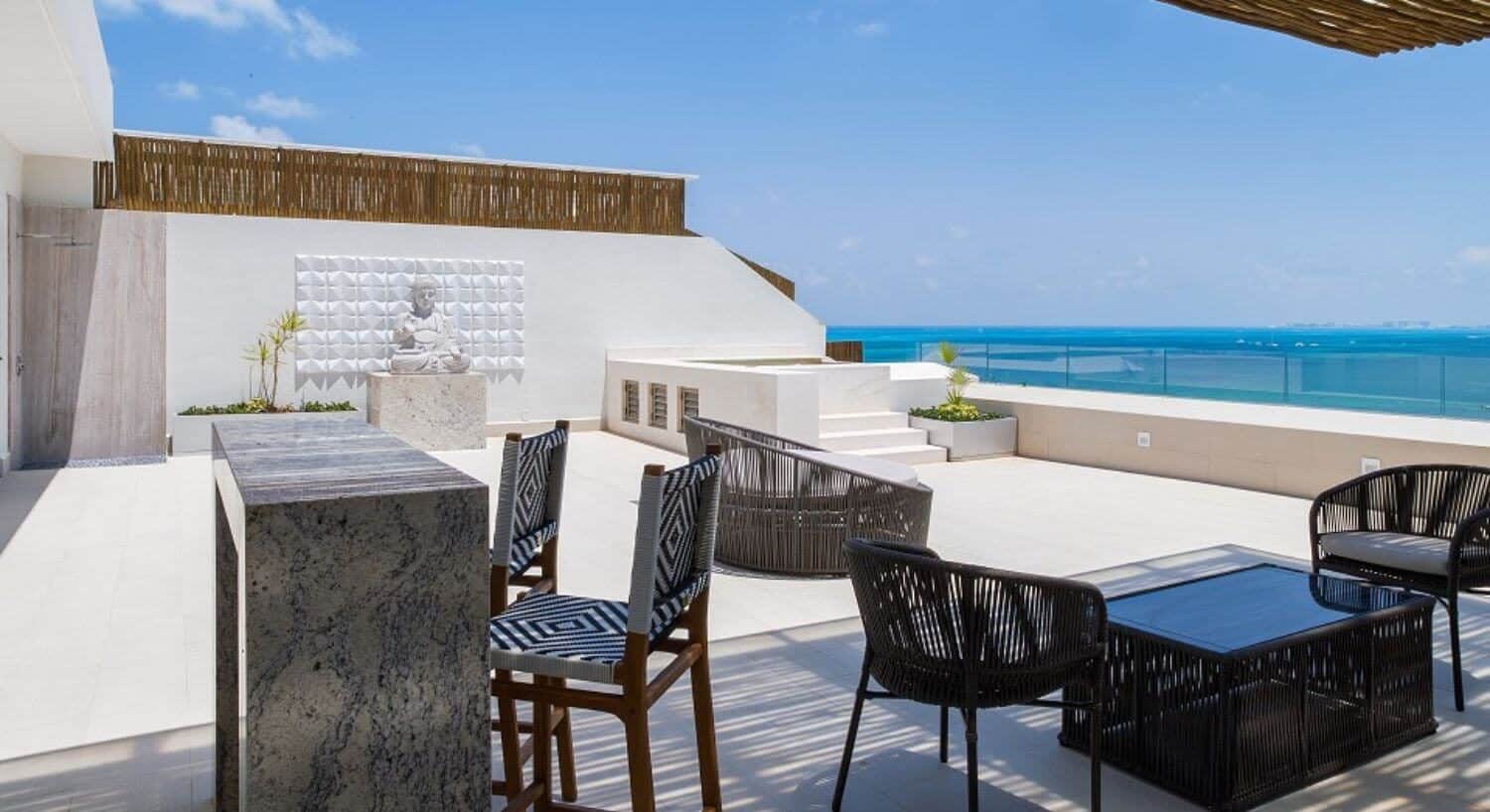 A rooftop terrace with a jacuzzi, bar with barstools, and patio furniture, with beautiful turquoise ocean views.