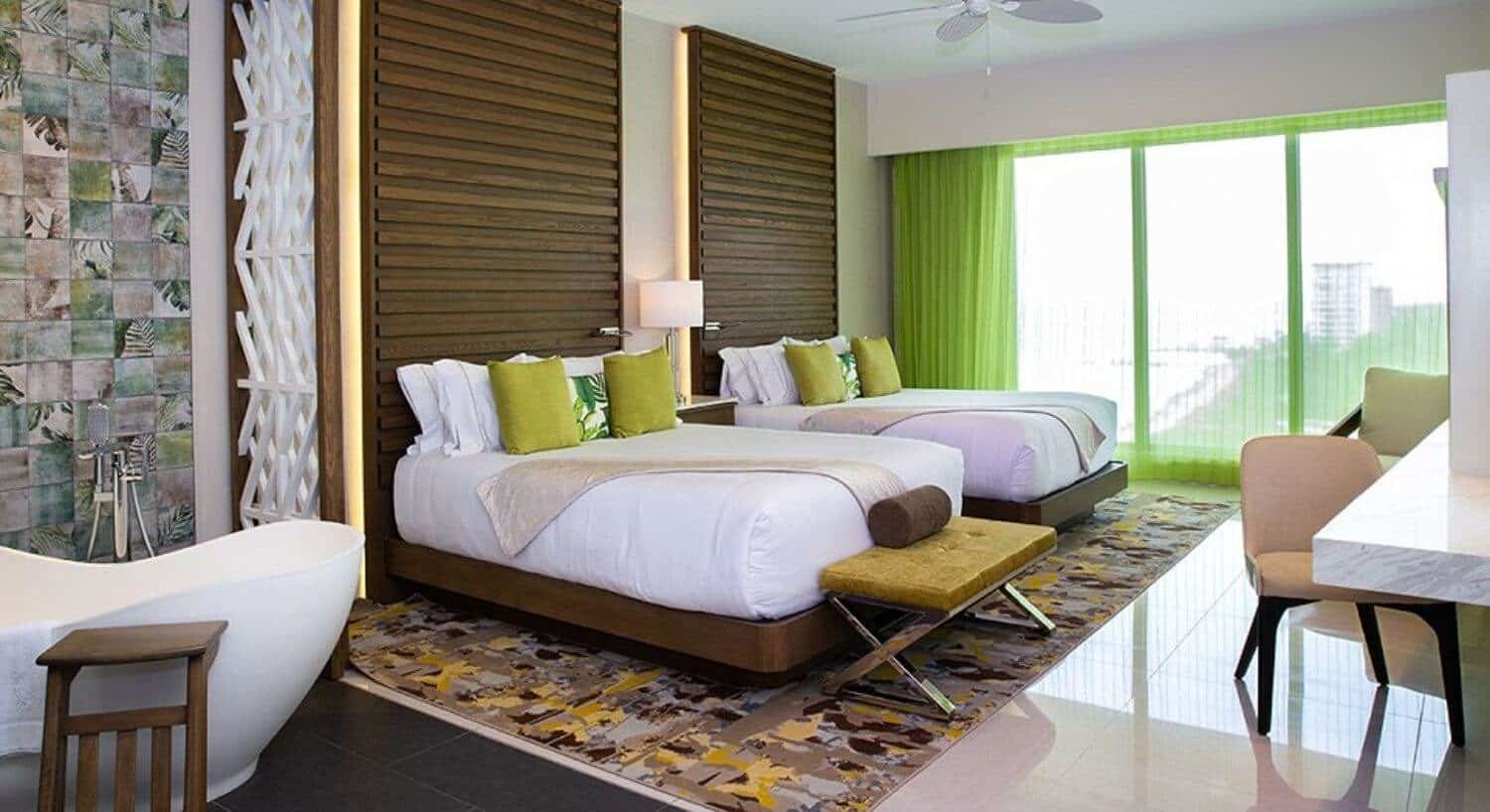 A bedroom with 2 queen beds with white, green and tan bedding, a large soaking tub, a writing desk and chair, and sliding doors out to a private balcony with sheer green curtains.