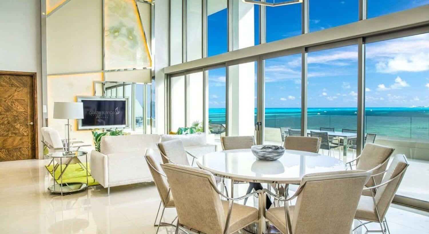 A large living and dining area with white sofa and chairs, flat screen TV, round dining table and plush tan chairs, and two story floor to ceiling windows, and sliding doors leading out to a private balcony with outdoor patio furniture, with stunning turquoise ocean views.
