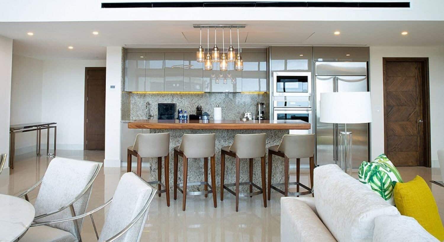 A gourmet kitchen with stainless steel appliances, a breakfast bar with four barstools, a dining table and chairs, and a plush sofa and floor lamp.