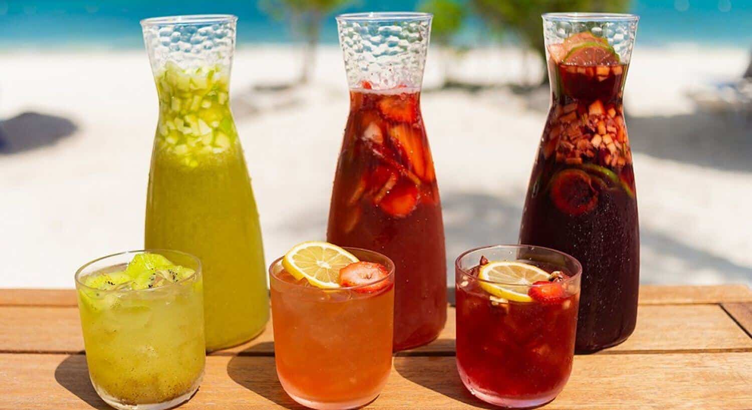 Three carafes and three glasses of various fruit sangrias in yellow, red, and purple.