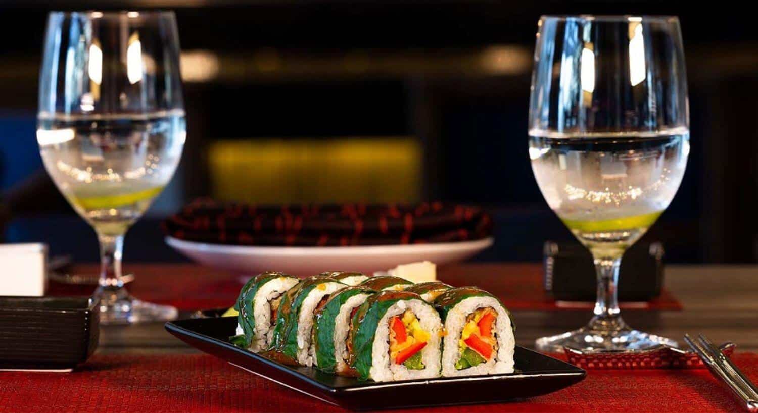 A black rectangular plate filled with 8 sushi rolls of cooked rice, fresh vegetables, and wrapped in seaweed, with 2 glasses of water next to it, on a table with red placemats.