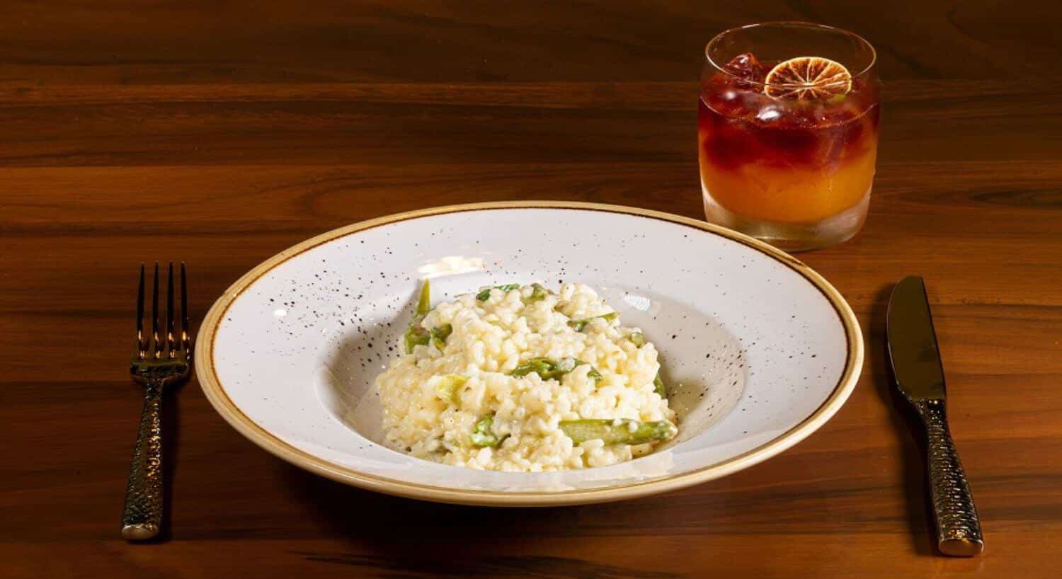 A white bowl of cooked white risotto with sprigs of cooked asparagus mixed in; along with silverware on either side, and a clear glass with an orange and brown cocktail with a dried lime slice on top.