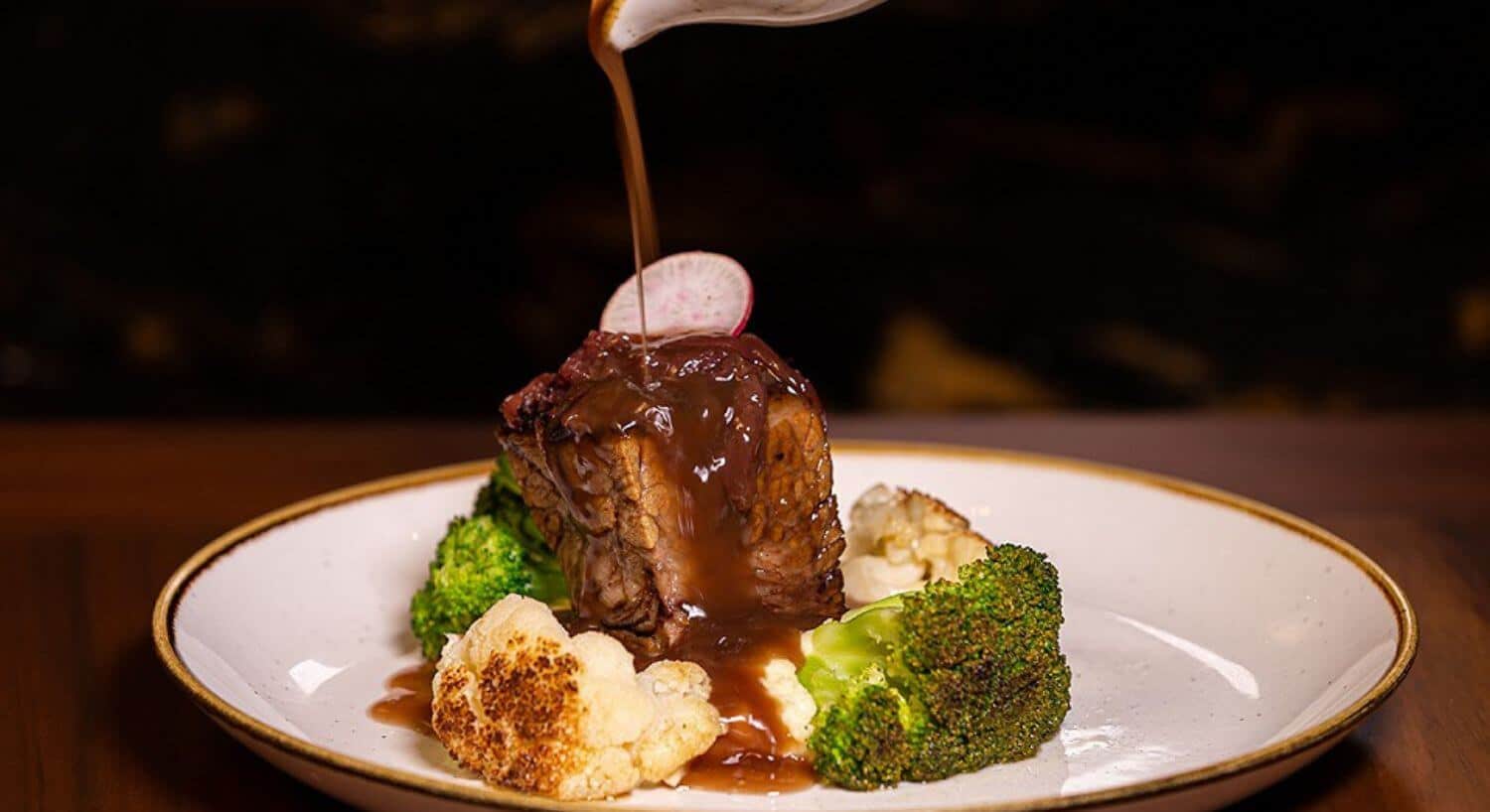 A white plate with a piece of filet steak atop a mound of mashed potatoes, with grilled broccoli and cauliflower pieces around it, and a brown gravy drizzled on the meat.