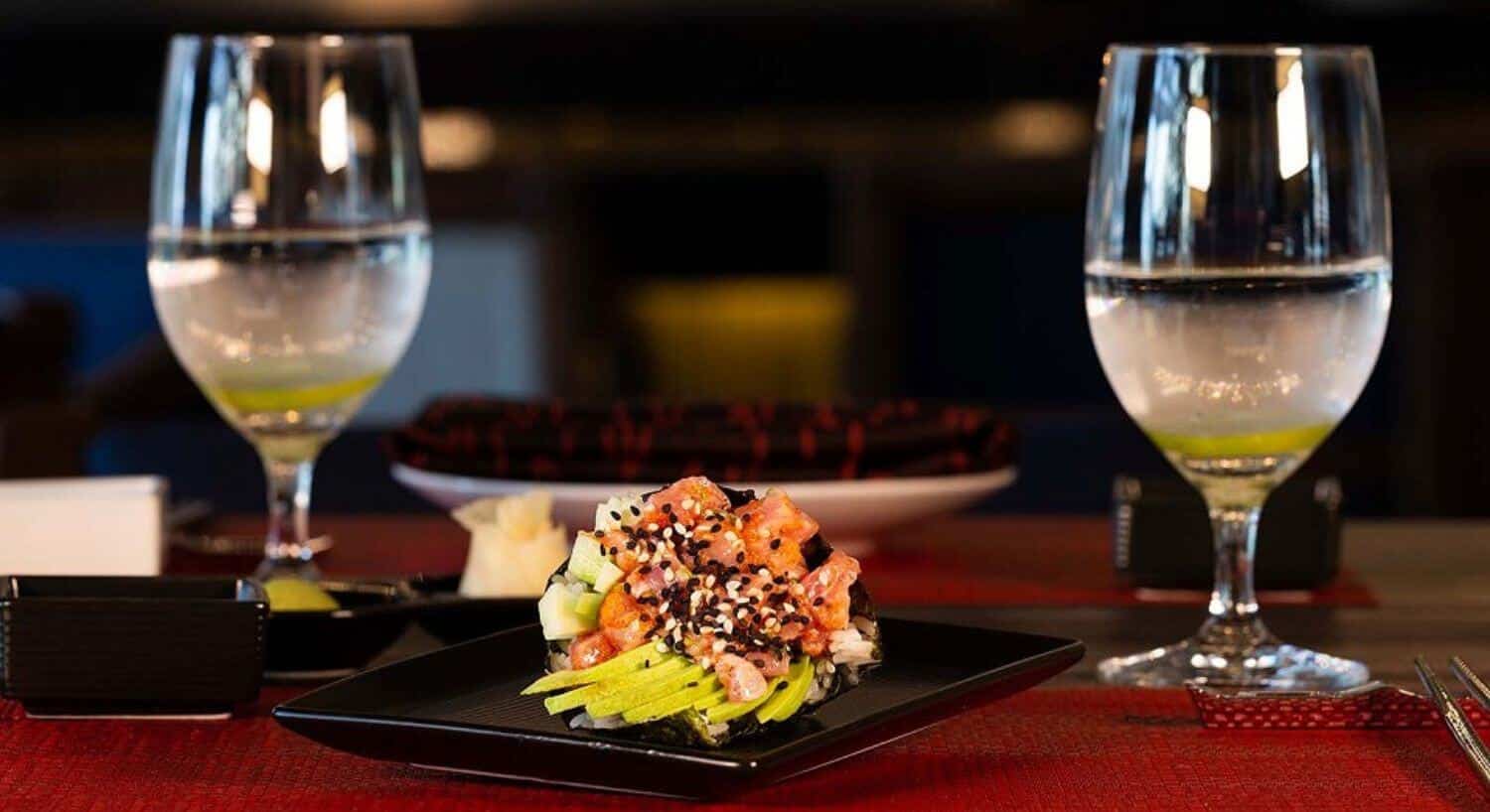 A square black plate on a red placemat with slices of avocado, tuna pieces, and white and black sesame seeds; with two glasses of ice water with lime slices.