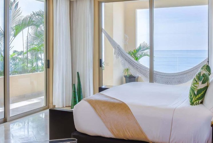 A bedroom at the corner of a building with a King bed with white, tan and green bedding, nightstands and lamps on either side of the bed, and sliding doors on the two corner walls opening out to a wrap around balcony with a balcony, lush palm trees and bushes, and ocean views.