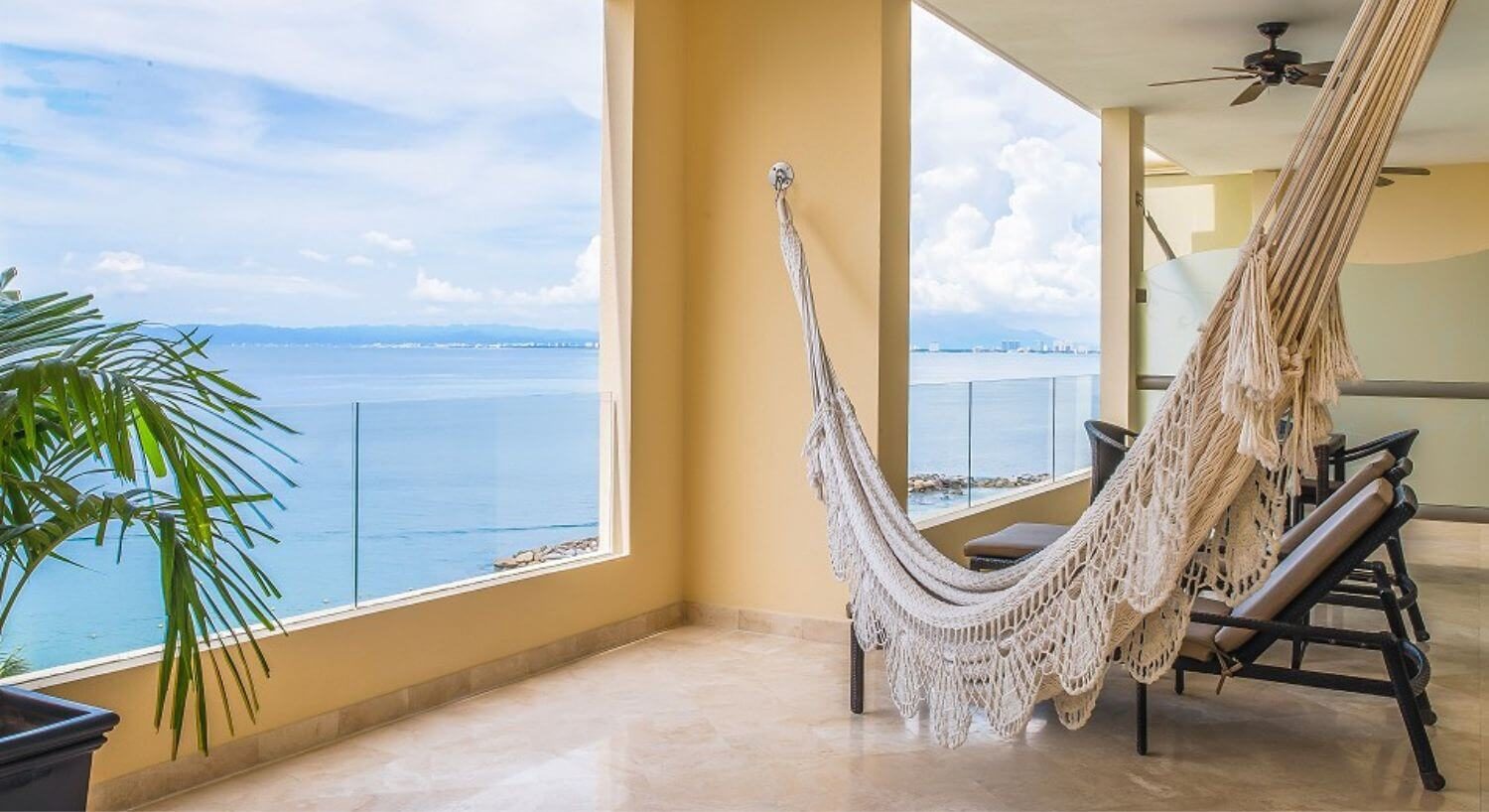A private outdoor balcony with 2 brown lounge chairs, an outdoor dining table and chairs, a hammock, and ocean views.