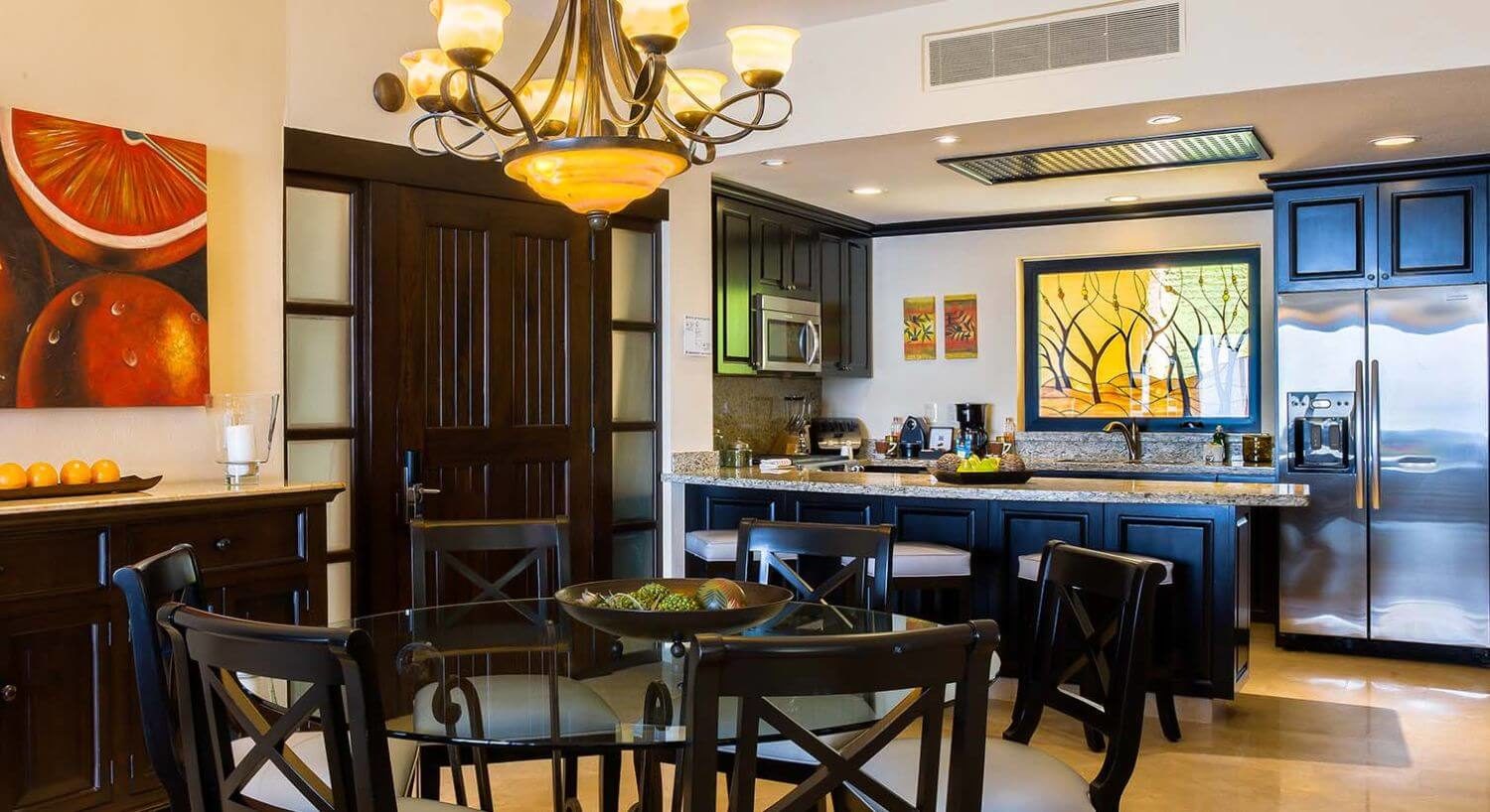 A kitchen and dining room with stainless steel appliances, granite countertops, rick dark brown furnishings, a glass tabletop, and a sideboard to match.