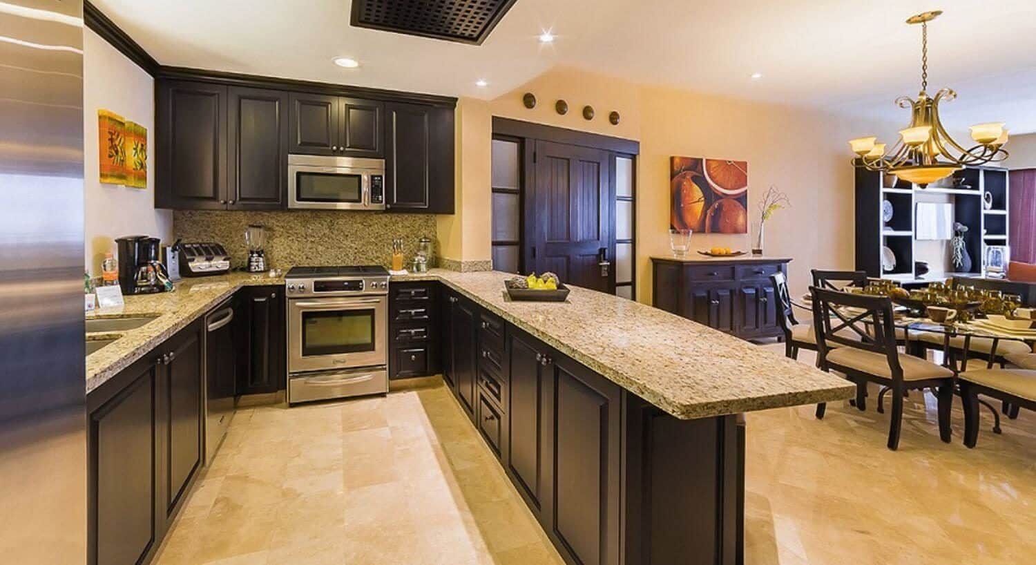 A U-shaped kitchen with stainless steel appliances, rich dark cupboards, granite countertops, a round glass dining table and plush chairs, a sideboard to match, and an entertainment center with flat-screen TV in the living room.
