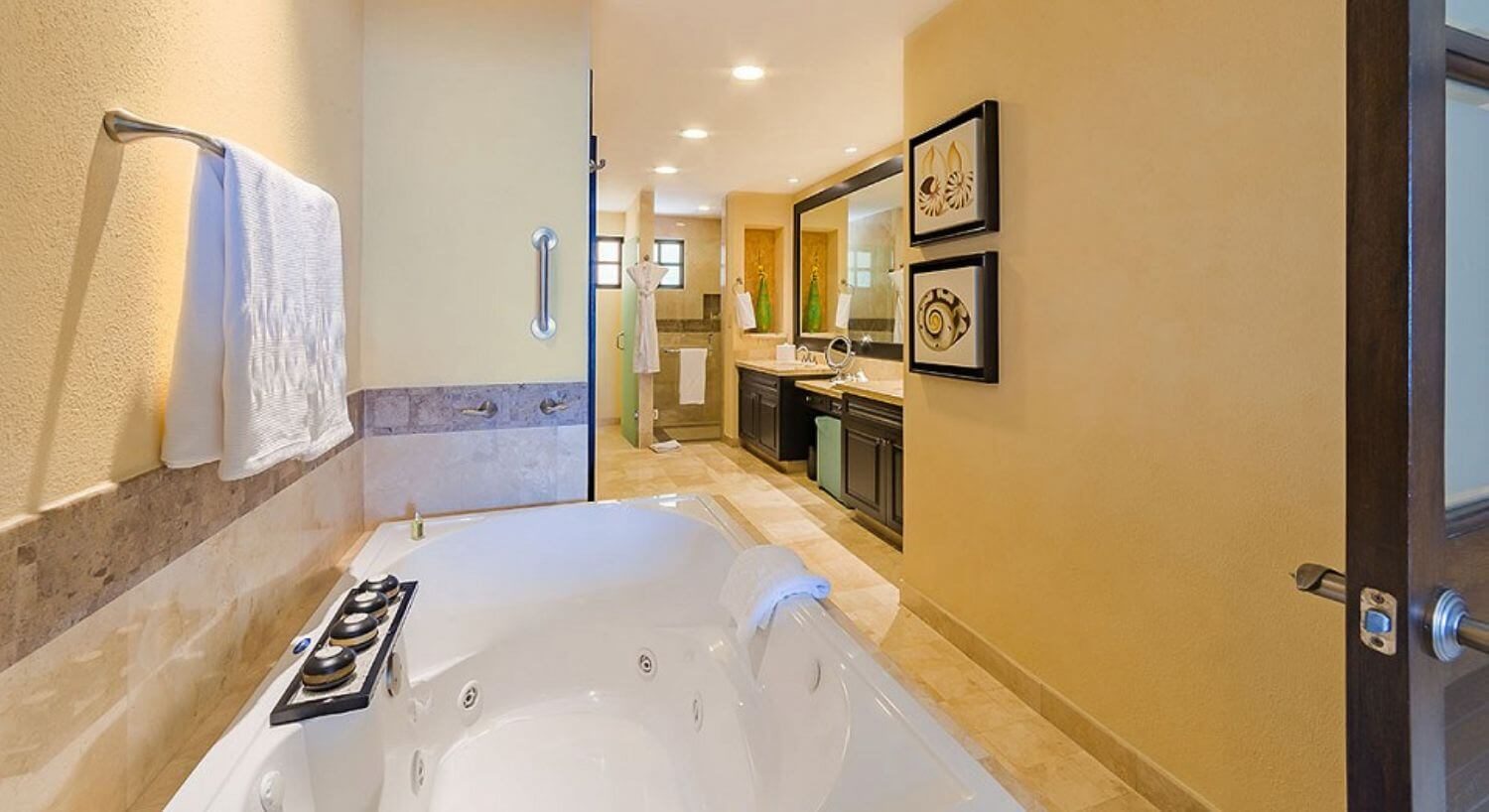 A large bathroom with a jetted bathtub, two granite bathroom sinks with a vanity with green ottoman in the middle, and a shower in the back.