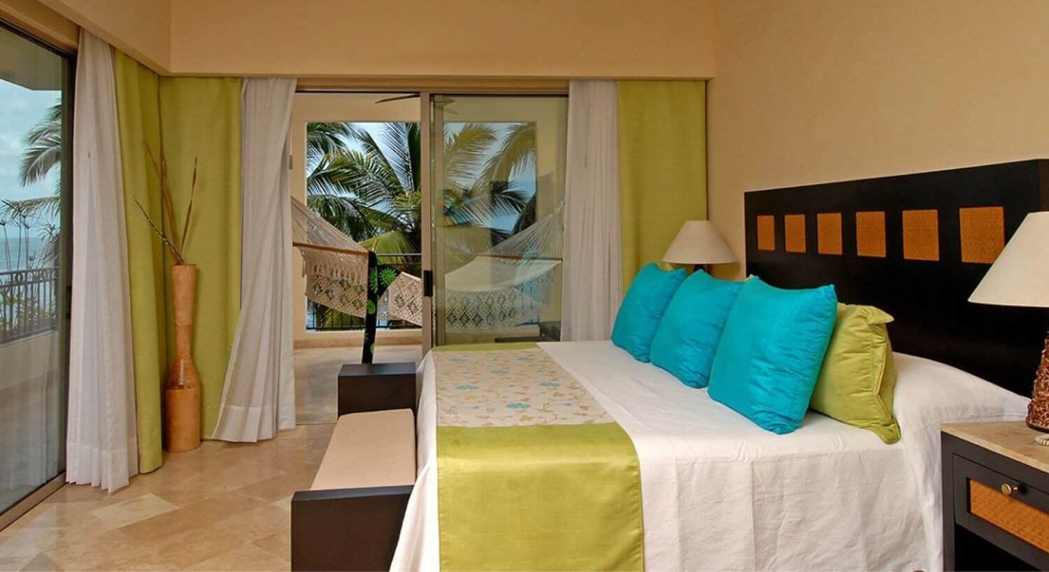 A bedroom with a king bed with white, lime and turquoise bedding, a sitting bench at the foot of the bed, lamps and nightstands on either side of the bed, and sliding doors on two sides of the room leading out to a wrap around balcony with an oversized hammock, and views of palm trees and the ocean.