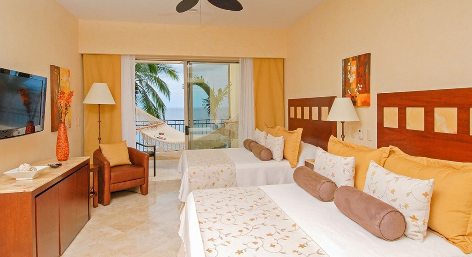 A bedroom with 2 queen beds with white and yellow bedding, a dresser and flat screen TV on the opposite wall, a plush leather armchair in the corner, and sliding glass doors leading out to a private balcony with oversized hammock, palm trees and ocean views.