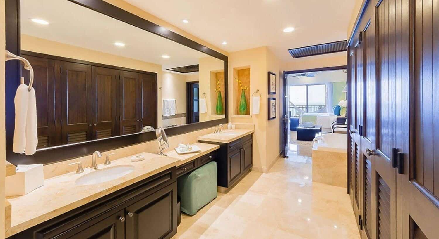 A spacious bathroom with double granite countertops and sinks, a vanity with a green ottoman in the middle, and jetted tub, with a bedroom with a loveseat, bed, and sliding doors leading out to a balcony with hammock.