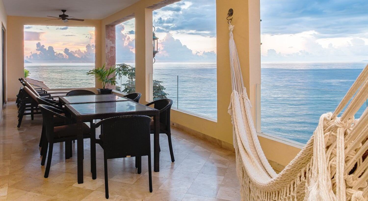 A private outdoor balcony with brown wicker patio furniture including tables and chairs, lounge chairs, and a hammock, with beautiful ocean views.