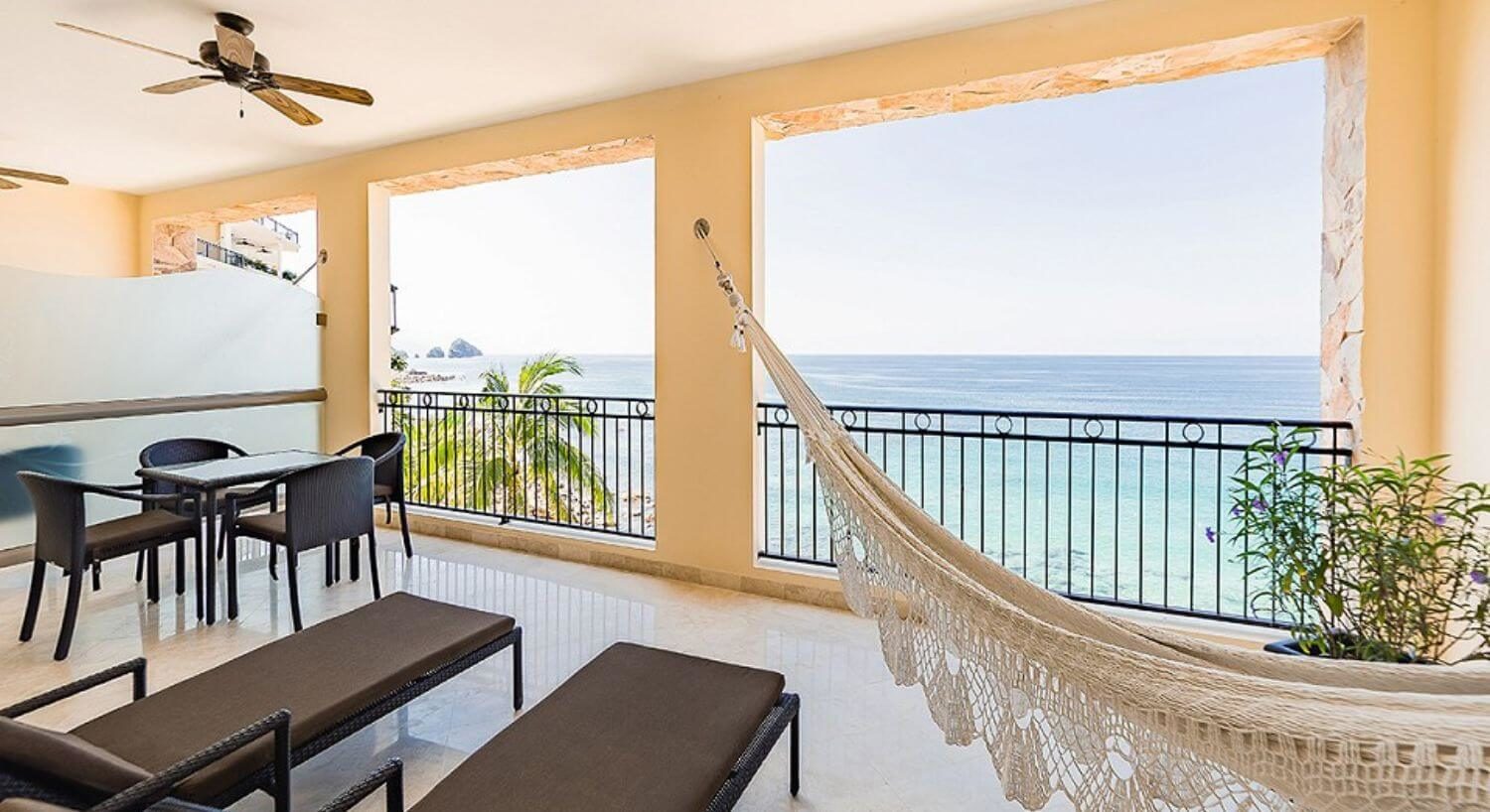 A private outdoor balcony with 2 brown lounge chairs, an outdoor dining table and chairs, a hammock, a wrought iron railing, and ocean views with the tops of palm trees popping up near the railing.