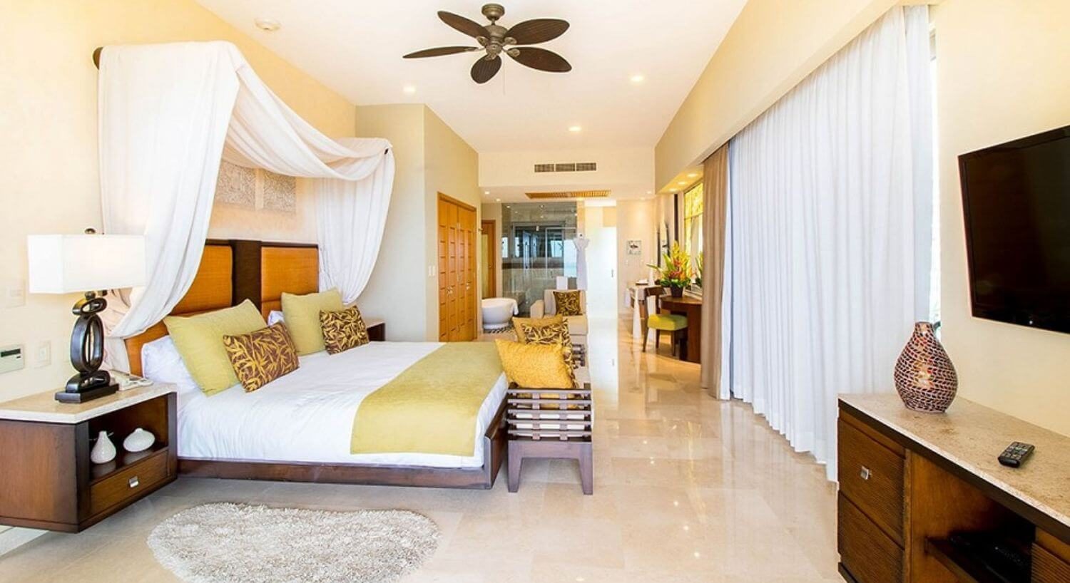 A spacious bedroom with a king bed with white, yellow and brown bedding, a white canopy draped over the head of the bed, nightstands and lamps on either side of the bed, a dresser and TV on the opposite wall, curtains covering sliding doors, and a bathroom with vanity, tub and shower.